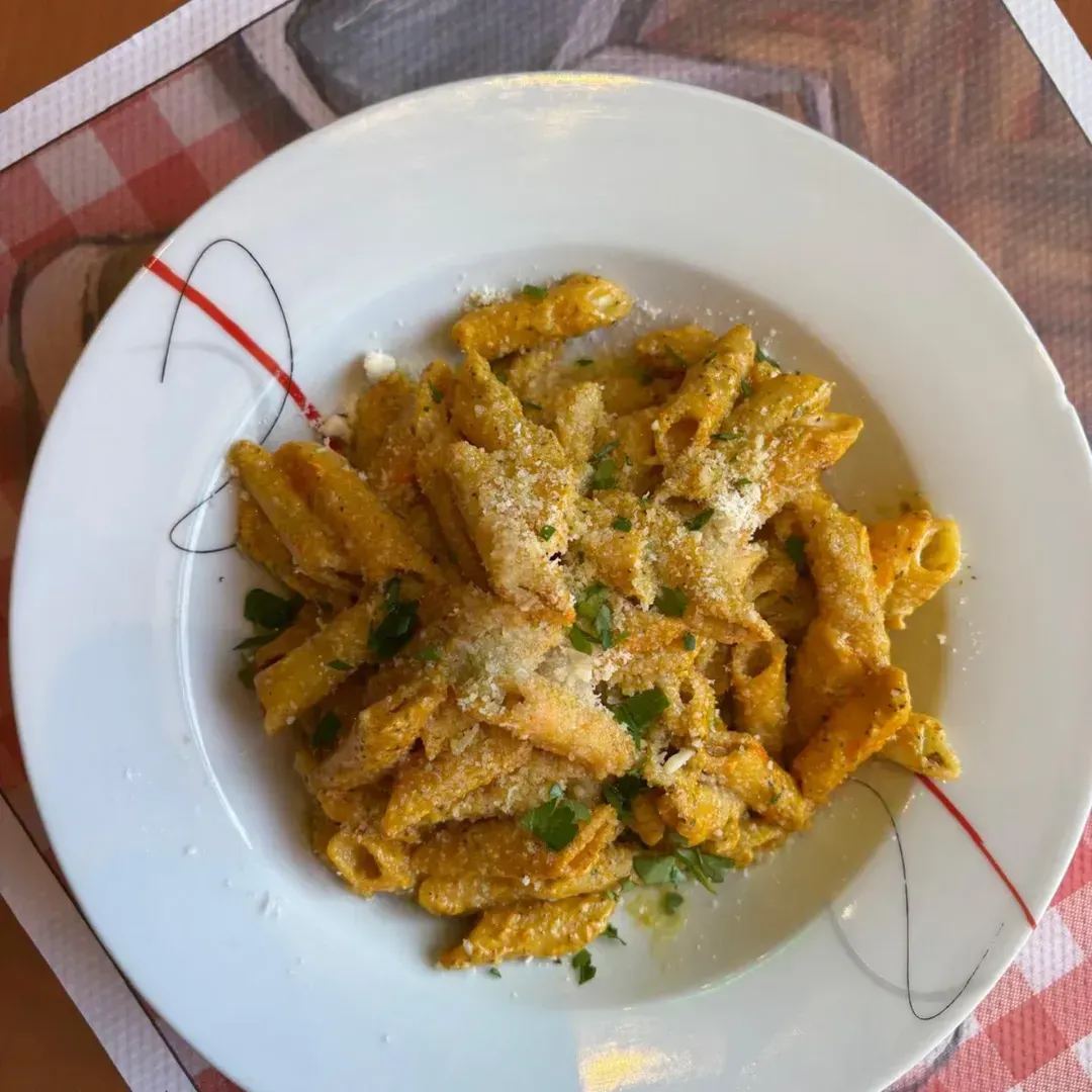 Un bol blanco lleno de pasta penne en una salsa cremosa de color naranja, cubierto con queso rallado y hierbas frescas.