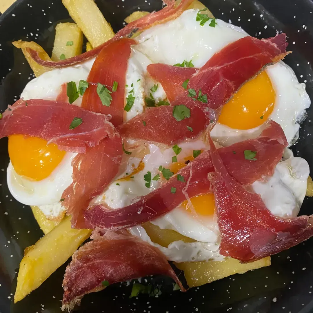 Huevos fritos con lonchas de jamón curado y patatas fritas, servidos en una sartén negra moteada.