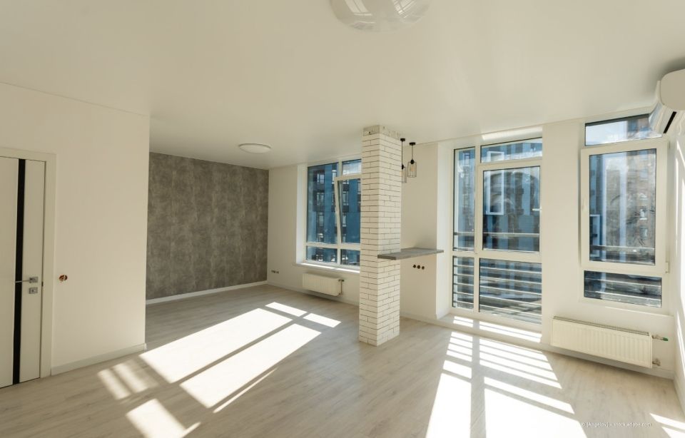 Helles, leeres, modernes Zimmer mit großen Fenstern, weißen Wänden und Sonnenlicht, das lange Schatten auf den Boden wirft.