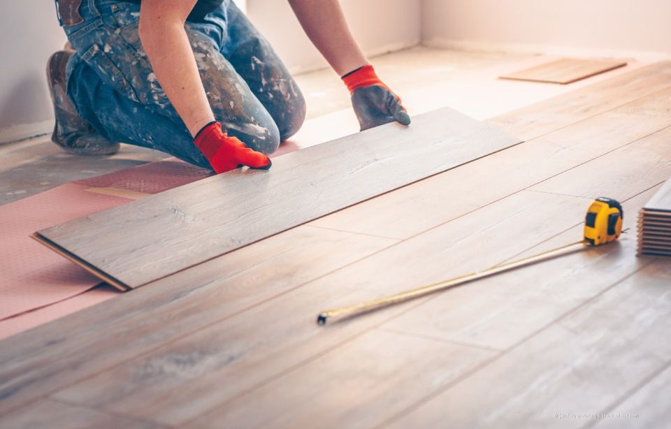 Eine Person verlegt in einem hellen Raum mit Maßband und roten Handschuhen einen hellen Holzboden.
