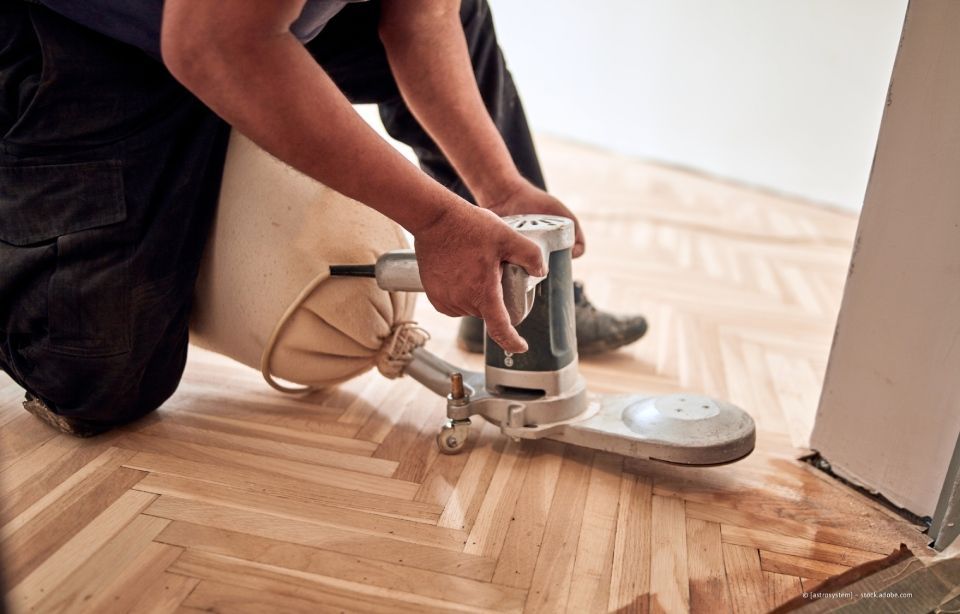 Eine Person schleift in einem renovierten Raum einen Holzboden mit einer Bodenschleifmaschine ab.