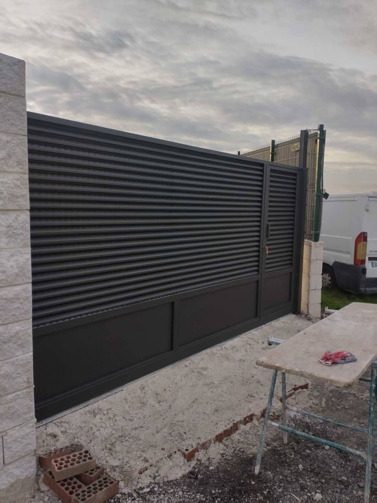 Portón de lamas horizontales de color gris oscuro con panel inferior macizo, junto a sección de lamas verticales a juego; base de hormigón y pared de ladrillo.