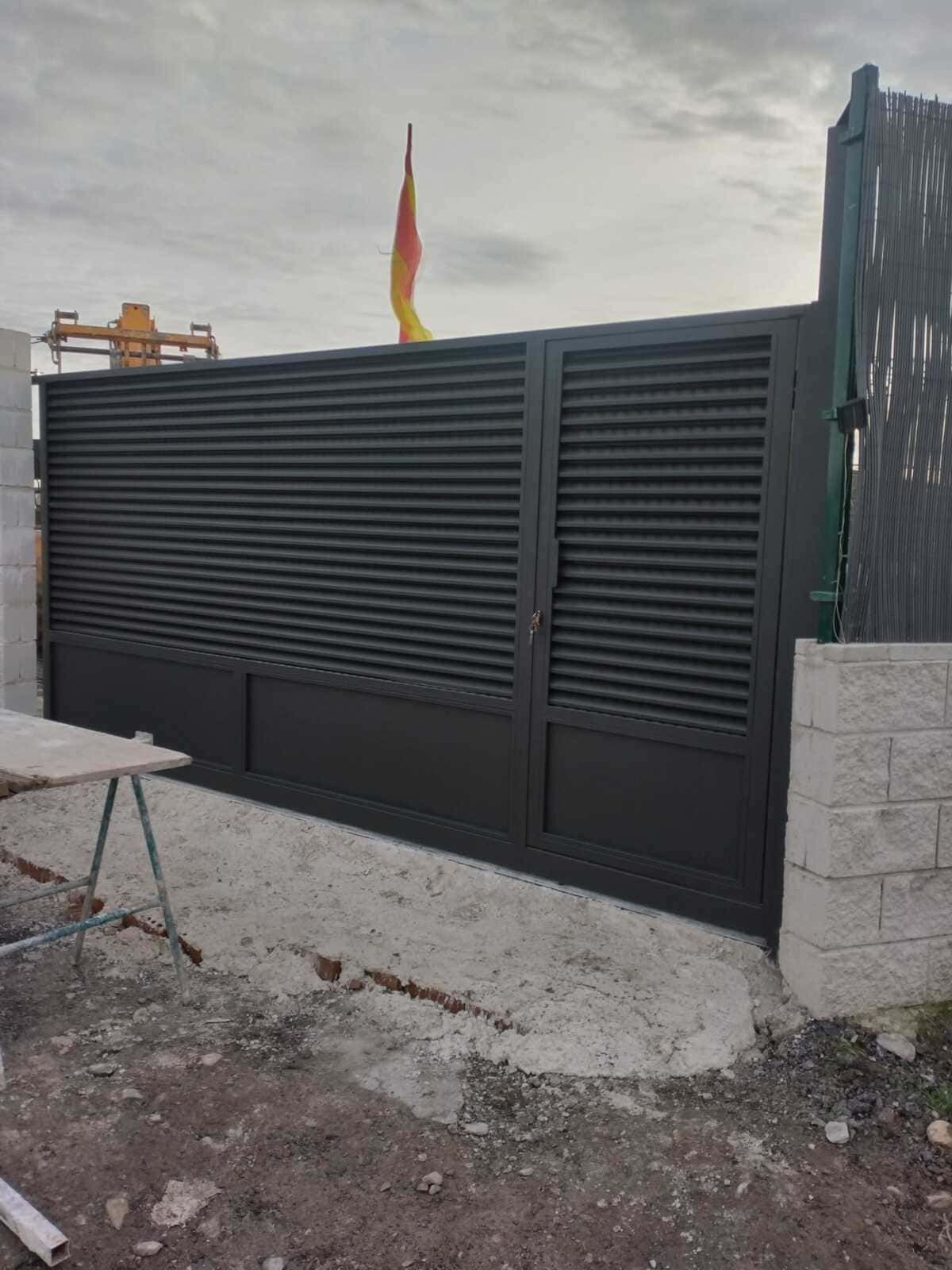 Portón de metal negro con lamas horizontales, base de hormigón, cielo nublado, bandera en la parte superior.