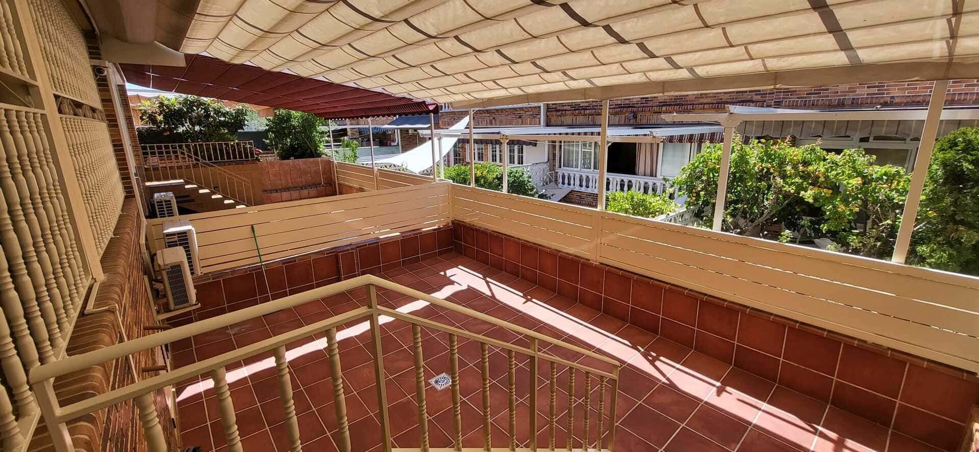 Patio exterior cubierto con barandillas de madera, suelo de baldosas y vegetación más allá del lado abierto.