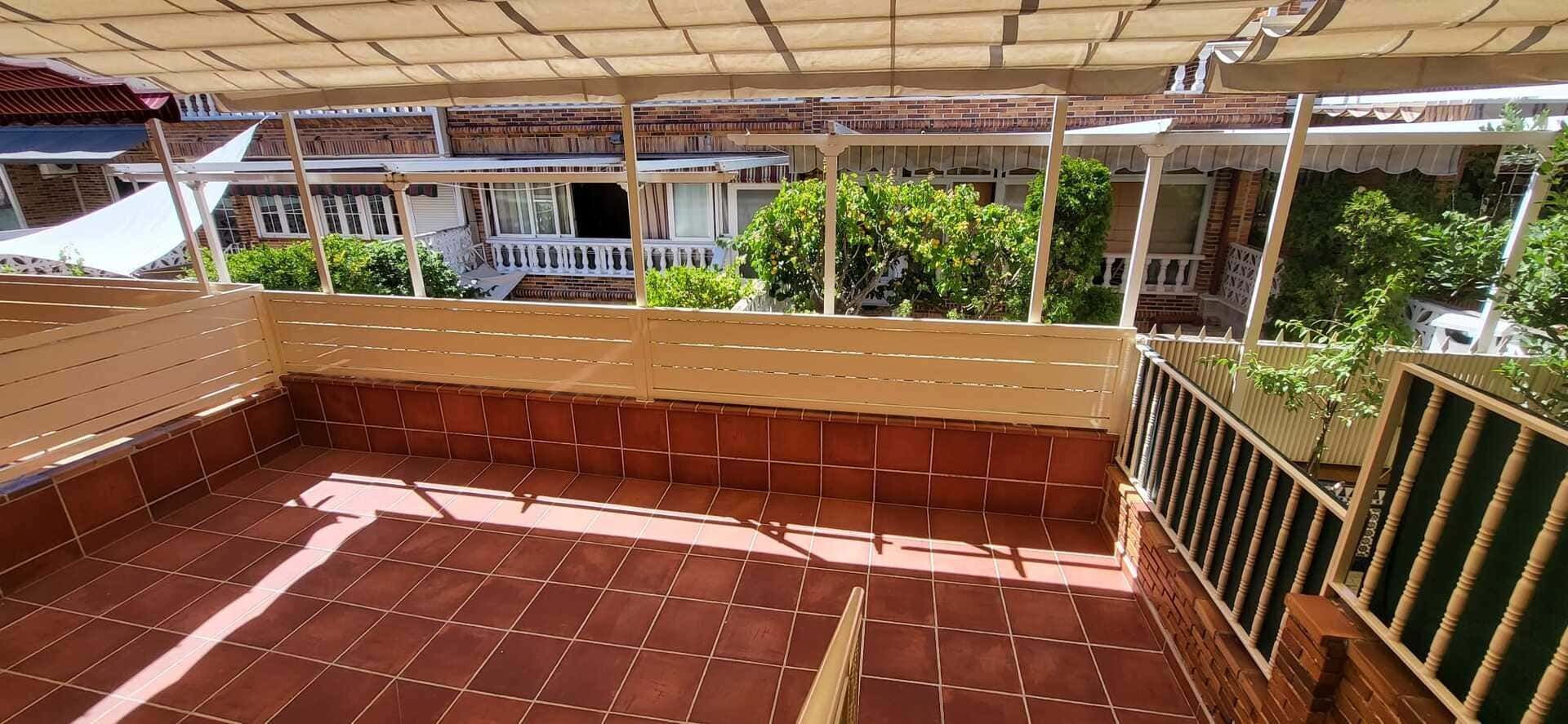 Terraza vacía con baldosas, barandilla marrón y toldo, con vistas a zonas verdes y edificios.