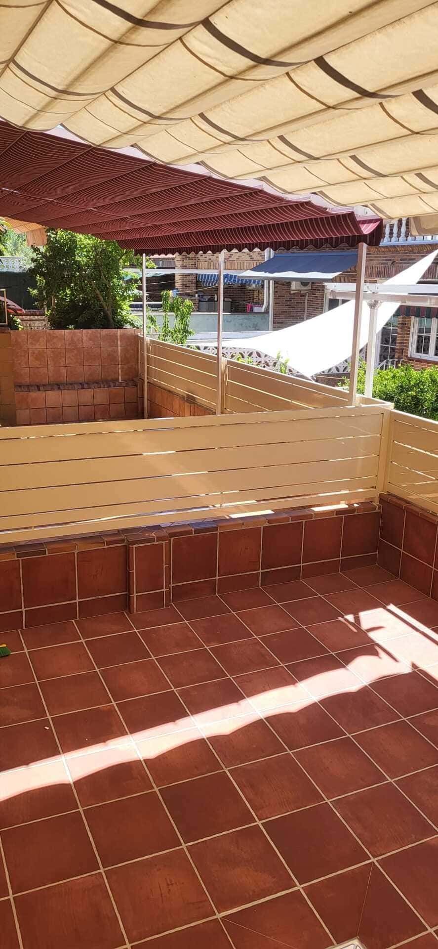 Patio exterior sombreado con suelo de baldosas, asientos de ladrillo y un toldo junto a la piscina.