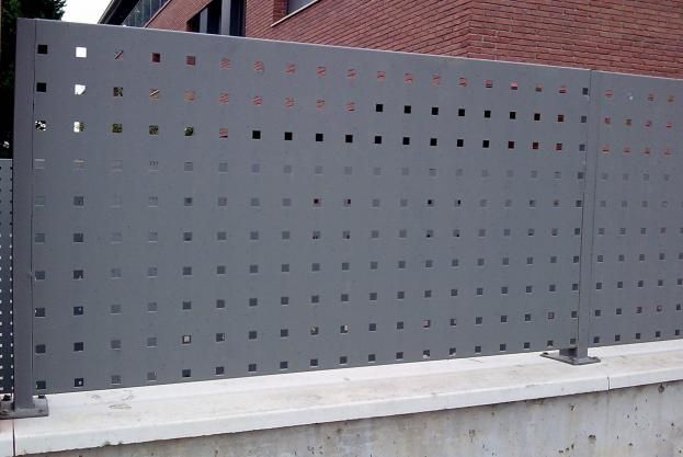 Una valla metálica gris con agujeros se encuentra delante de un edificio de ladrillos.