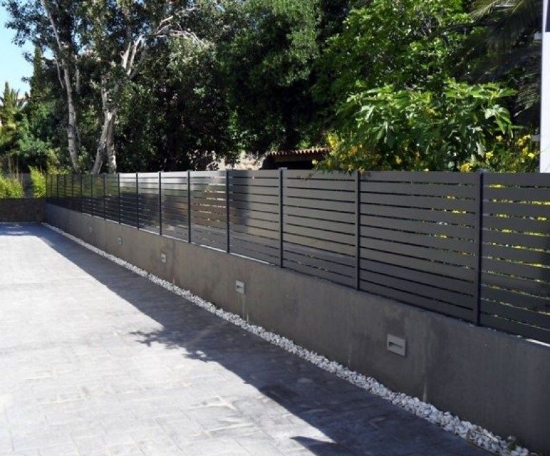 Una valla negra rodea un estacionamiento con árboles al fondo.