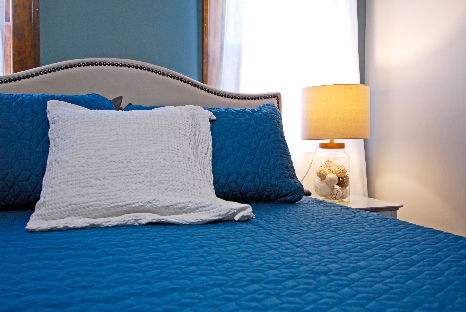 A bed with blue sheets and white pillows in a bedroom with a lamp.