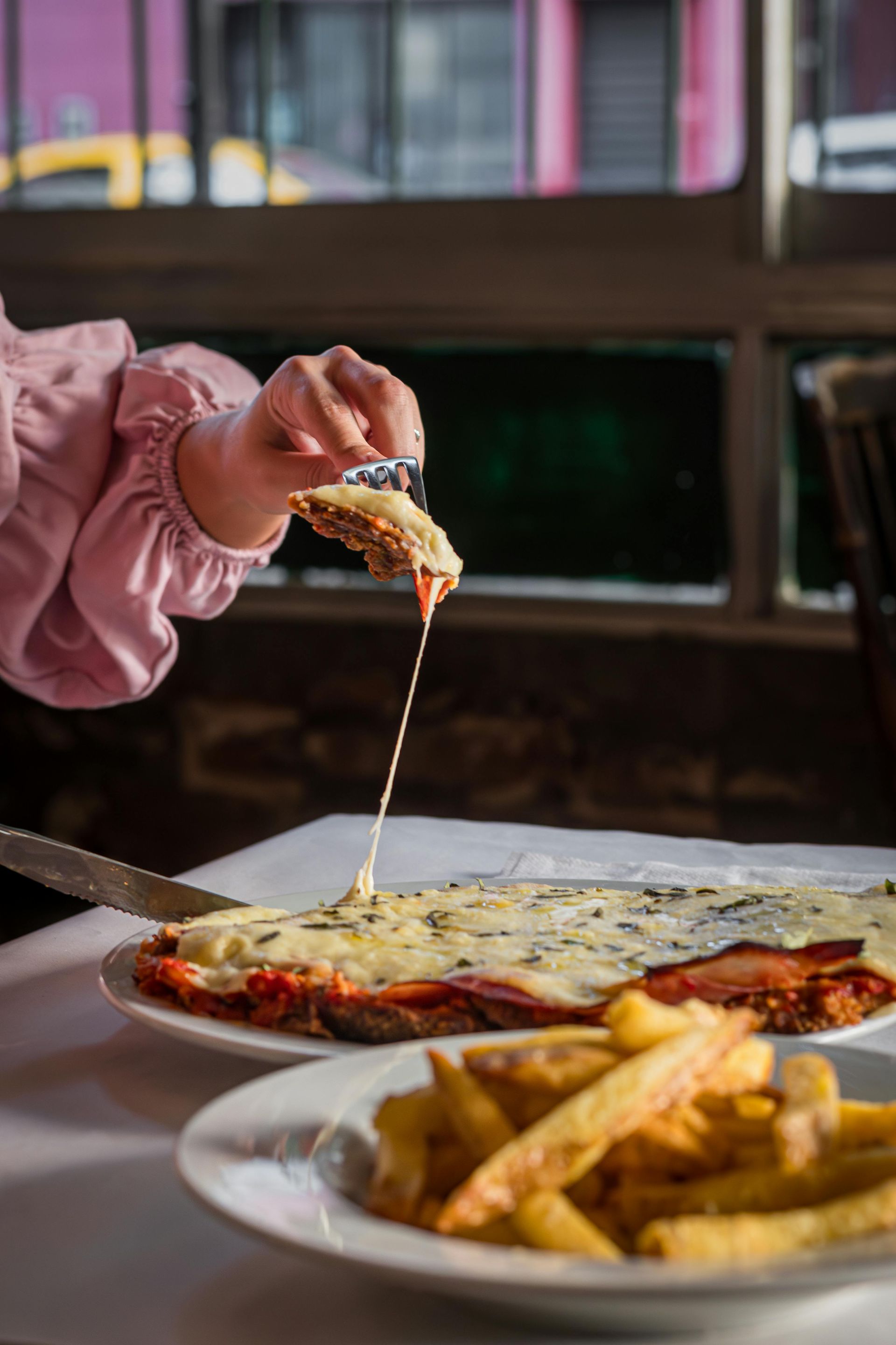 La mano de una persona levanta una porción de pizza con queso sobre un plato, con papas fritas en primer plano.