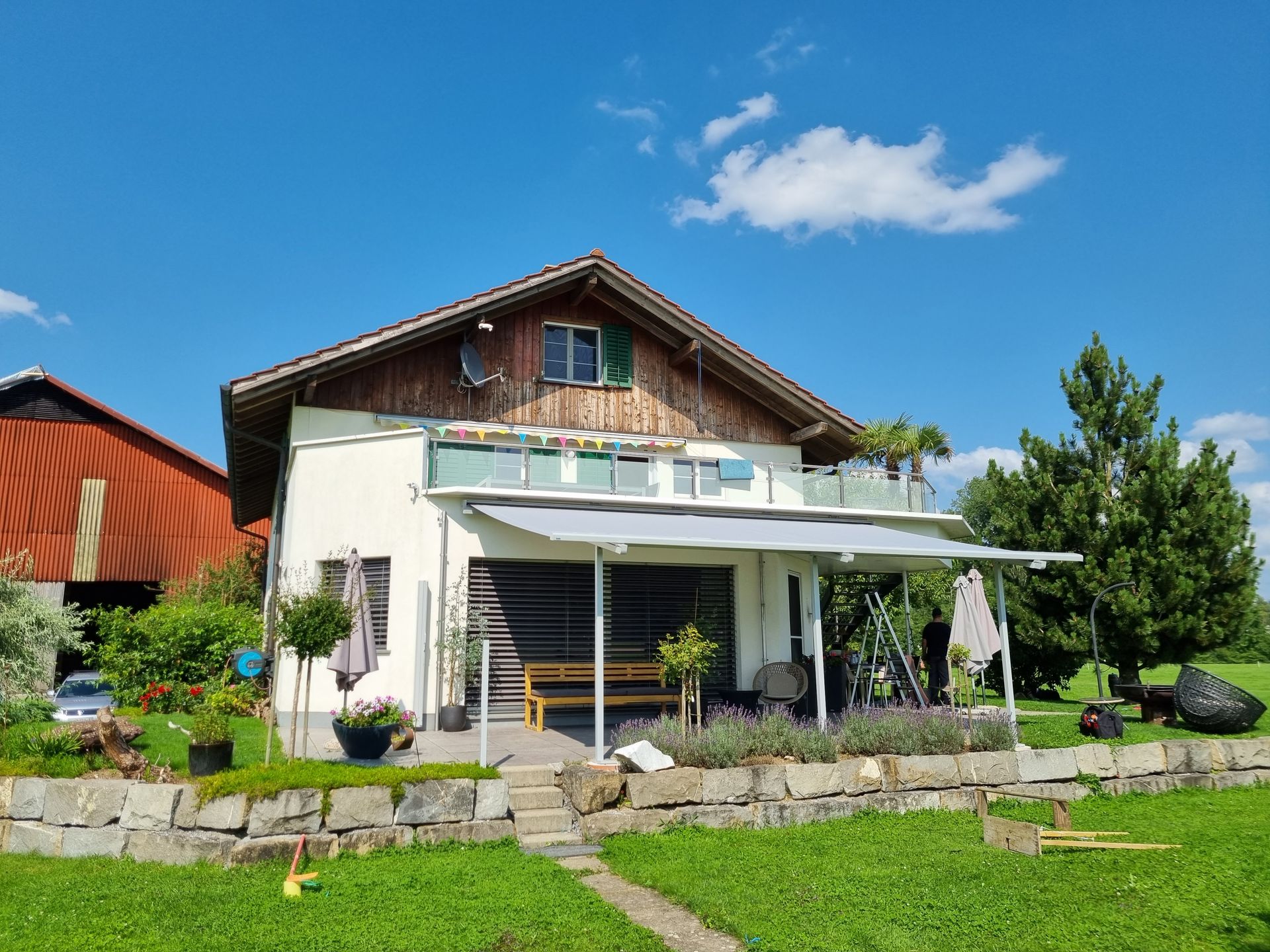 Weisses Haus mit braunem Dach und Balkon, in einem grünen Hof unter blauem Himmel.