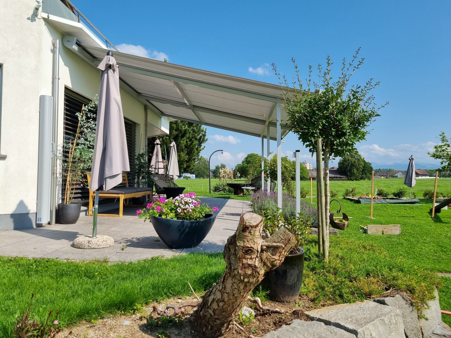 Eine Terrasse mit Baldachin bietet Blick auf einen grünen Hof mit Blumen, einem Baum und blauem Himmel.