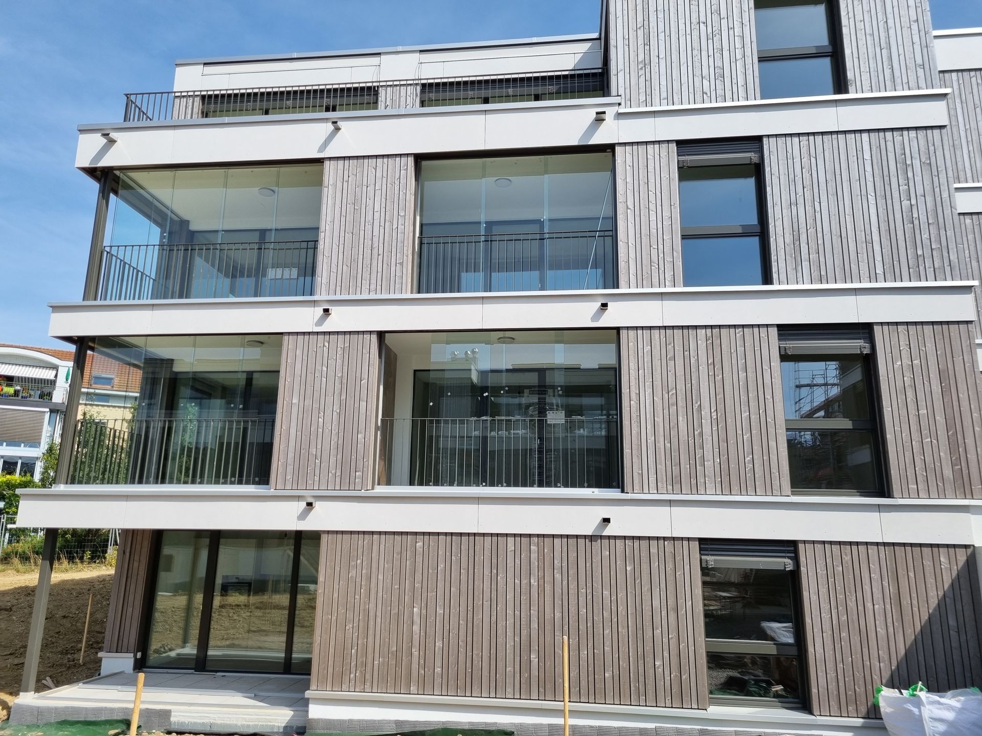 Mehrstöckiges, modernes Gebäude mit Holzfassade, Balkonen und grossen Fenstern; sonnige Außenanlage.