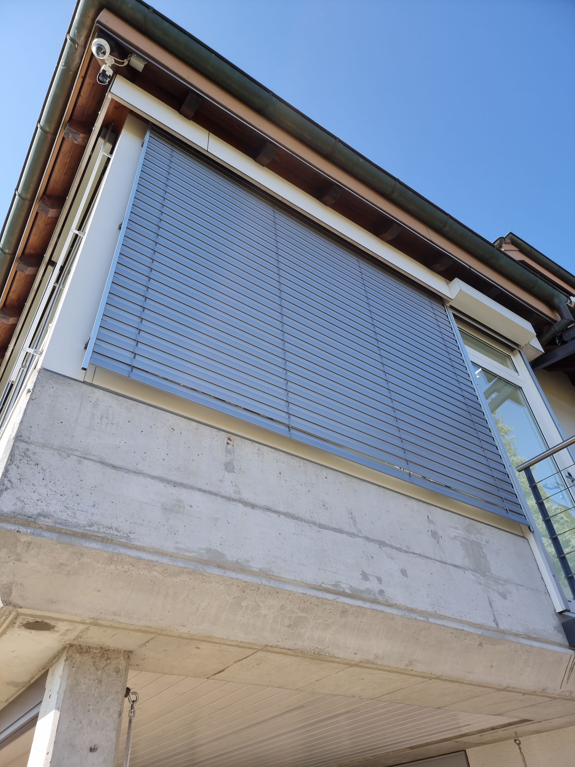 Aussenansicht eines Gebäudes mit Betonsockel, grossem Fensterrollo und klarem blauen Himmel.
