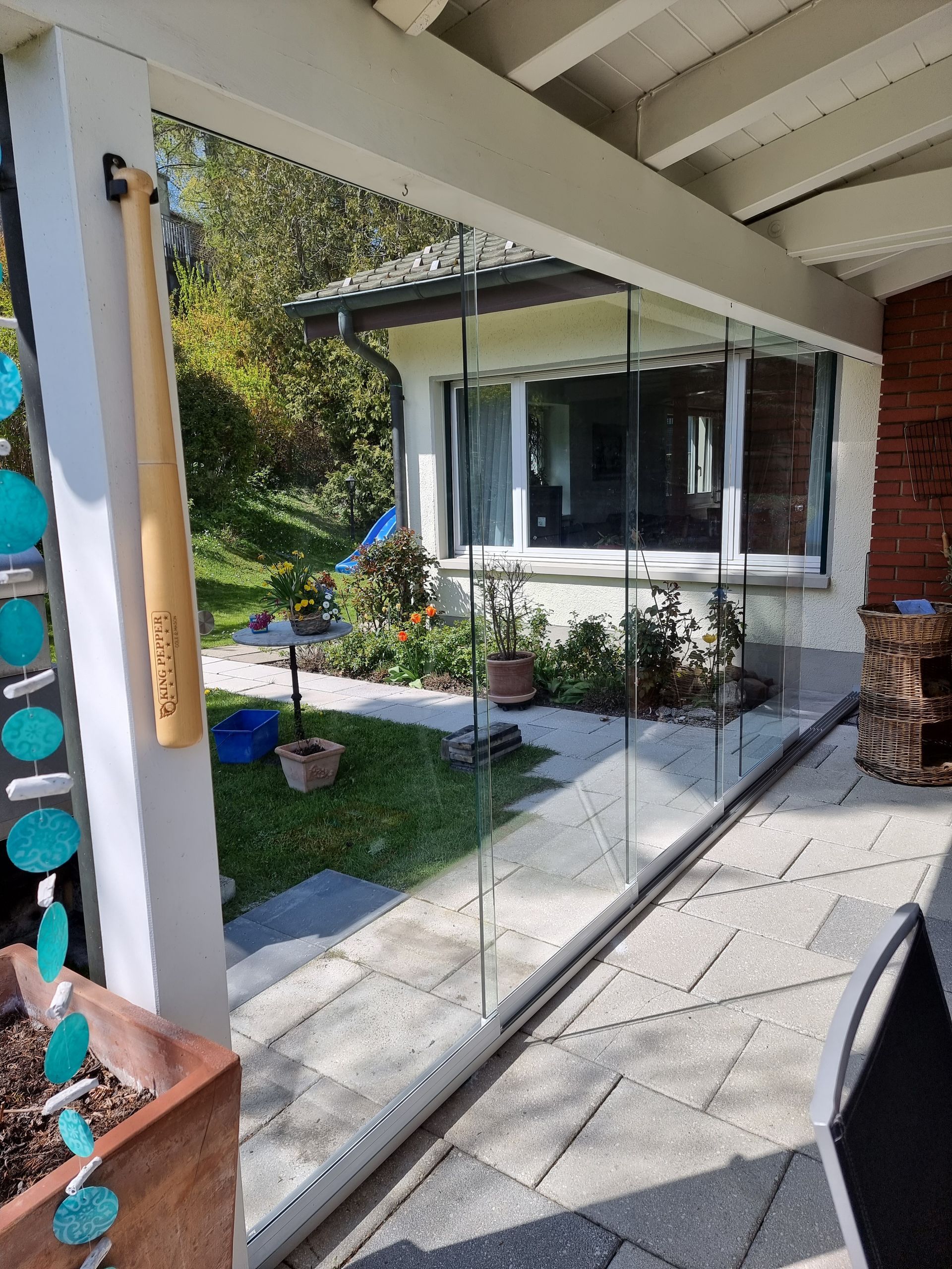 Terrasse mit Glaswänden und Blick auf einen Garten mit Blumen, Bäumen und einem Haus.