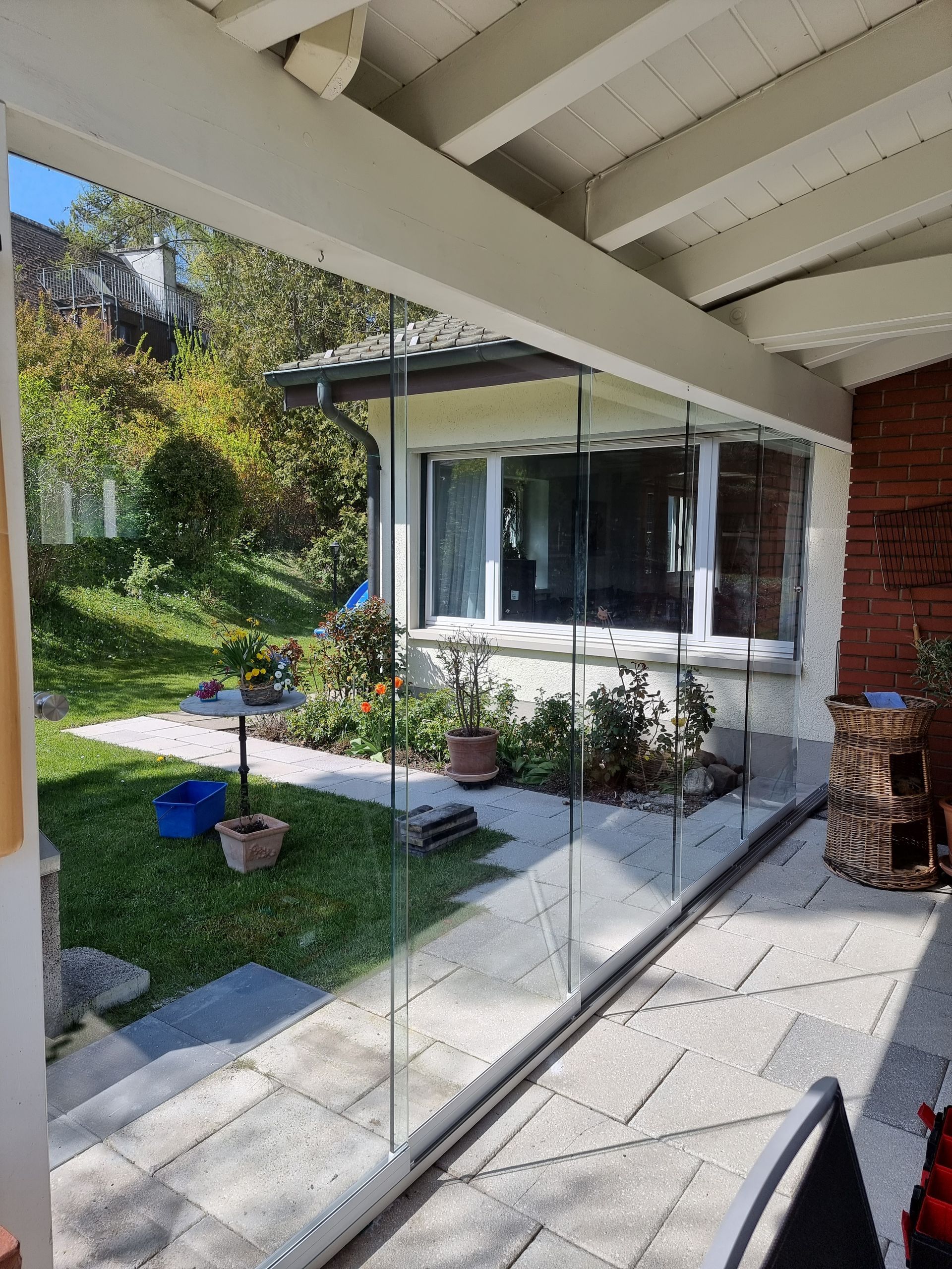 Terrasse mit Glaswänden und Steinboden, mit Blick auf Garten und Haus. Helles Tageslicht.