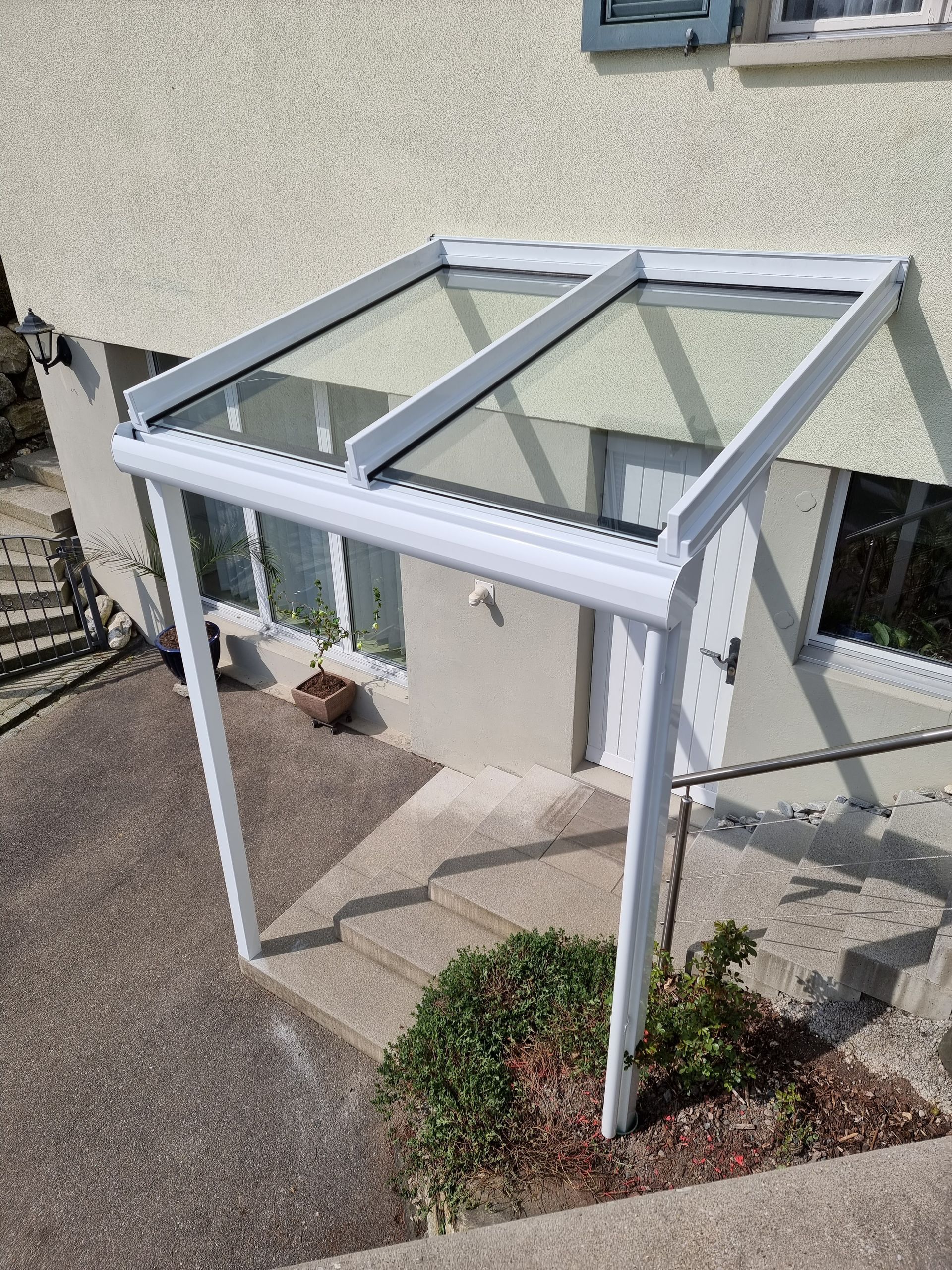 Weissgerahmtes Glasdach über einem Eingang, zu dem Stufen führen; helle Gebäudeaussenseite.