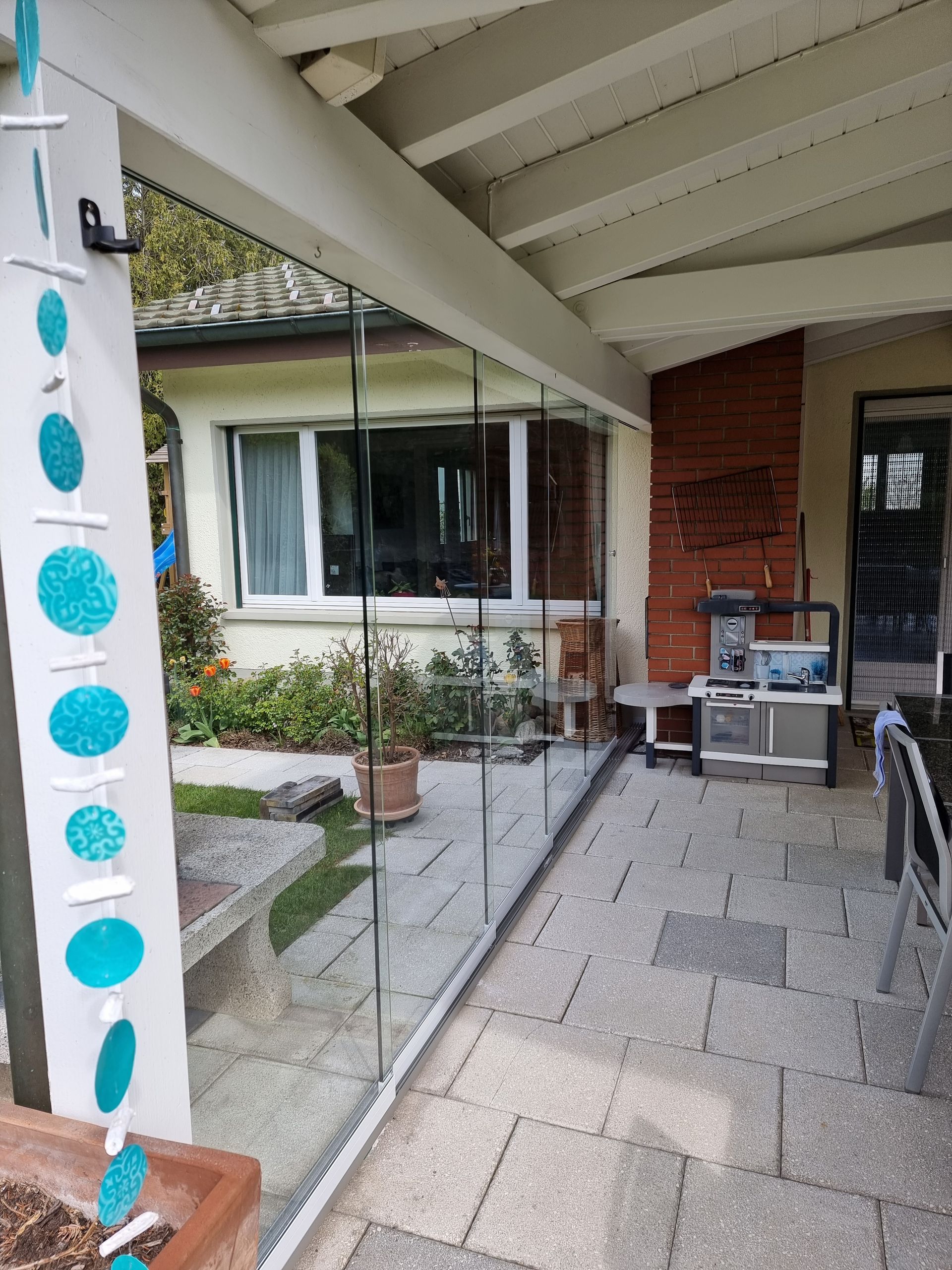 Terrasse mit Glaswänden, einem Haus im Hintergrund und hängenden türkisfarbenen Dekorationen.