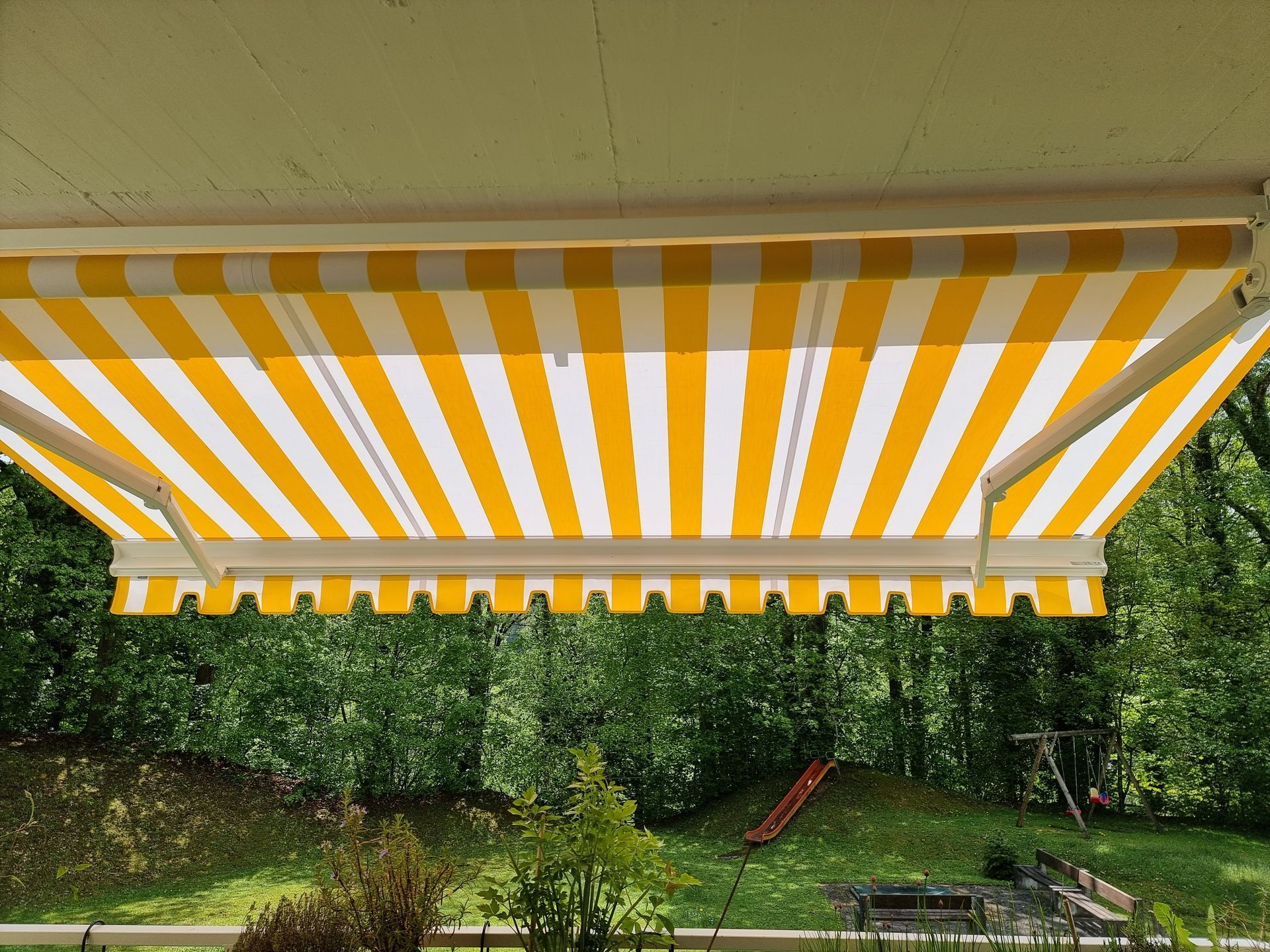 Gelbweiss gestreifte Markise über einem Hinterhof mit Rutsche und Bäumen.