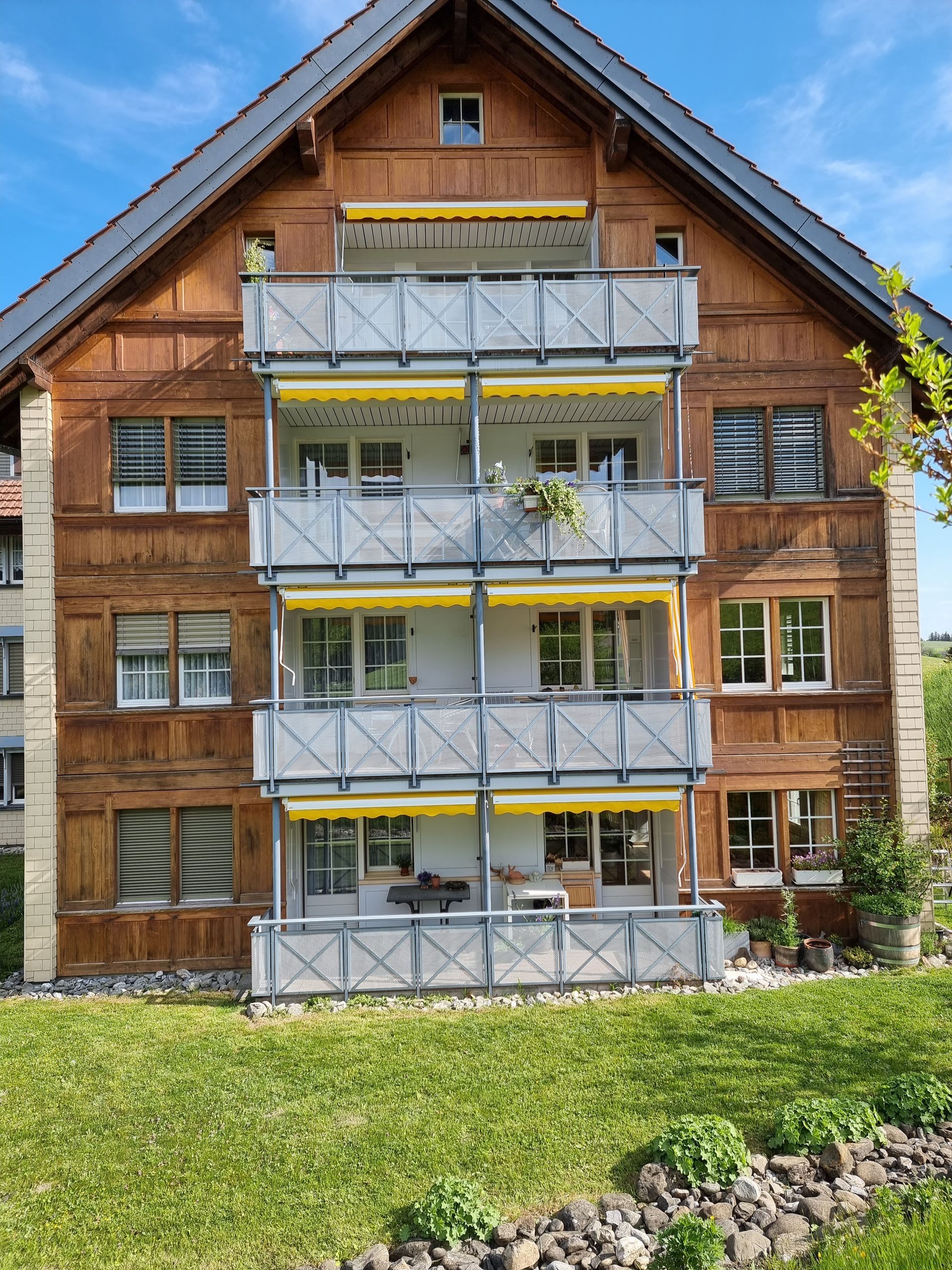 Dreistöckiges Gebäude aus Holz und Stuck, mit Balkonen, gelben Markisen und einem Rasenhof.