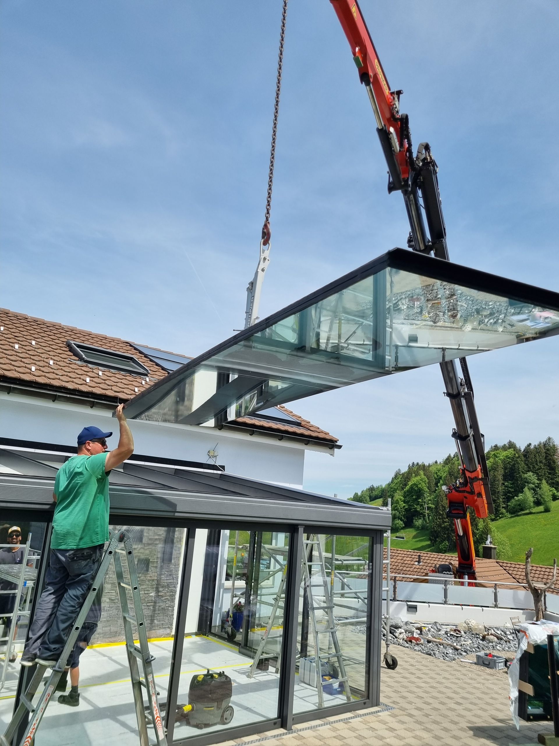 Ein Kran hebt eine Glasdachplatte auf ein Haus, ein Arbeiter auf einer Leiter führt die Platte.