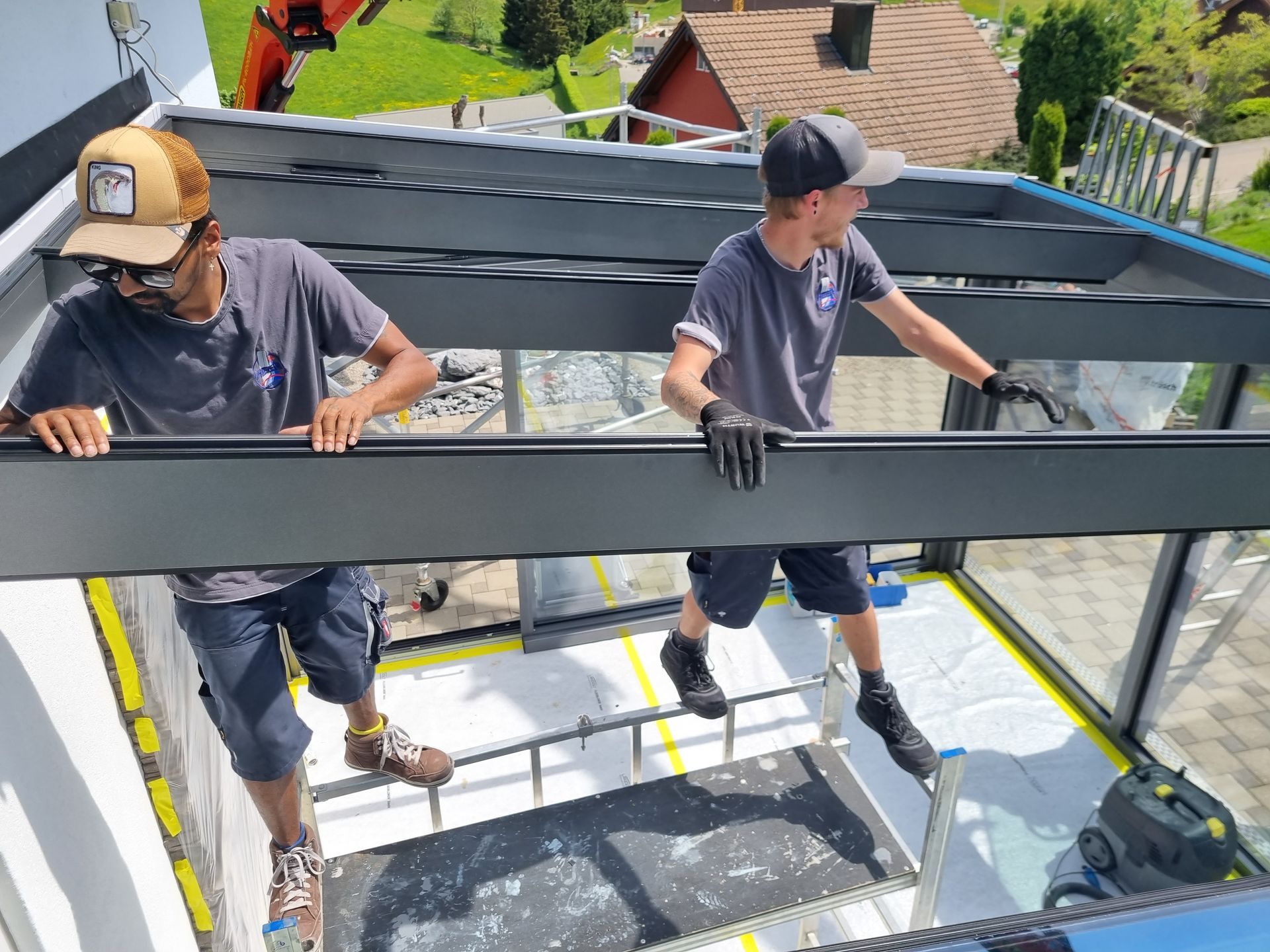 Zwei Arbeiter montieren ein Glasdach. Einer trägt einen Hut, der andere Handschuhe. Aussenaufnahme bei Tag.