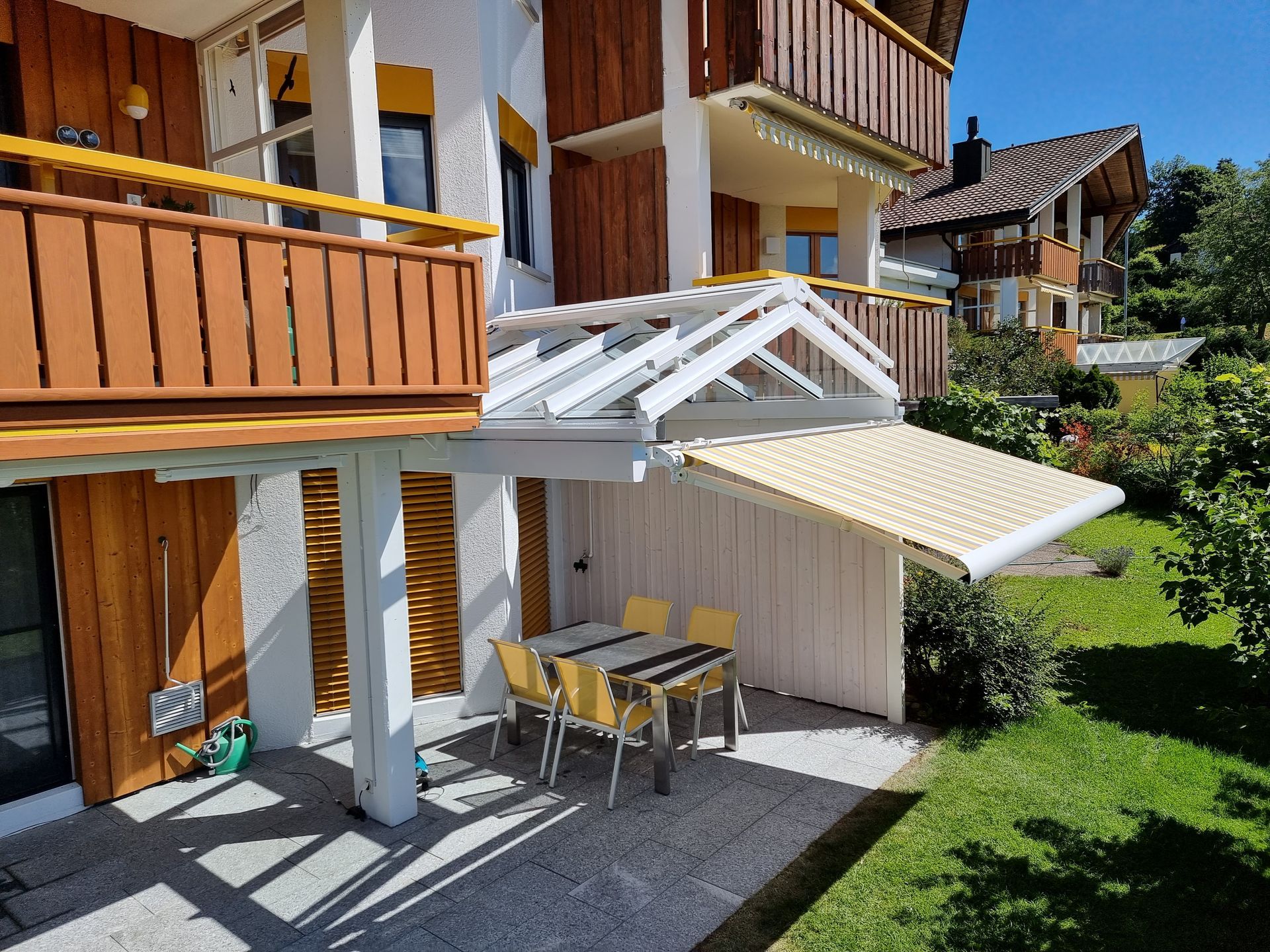 Ein mehrstöckiges Gebäude mit Holzbalkonen, Essbereich im Freien und einem Rasenhof an einem sonnigen Tag.