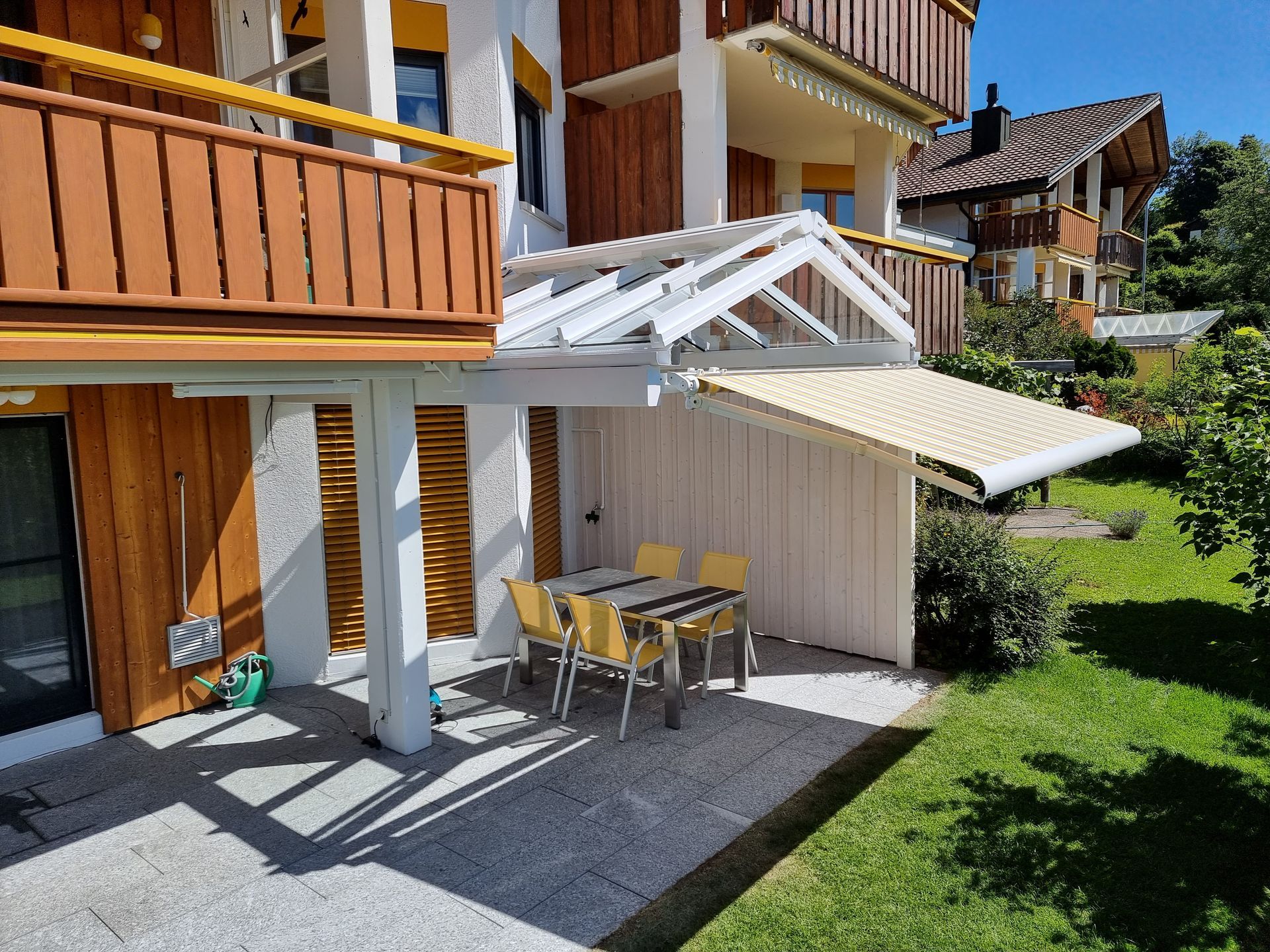 Aussenterrasse mit Tisch und Stühlen unter einer einziehbaren Markise neben einem Holzgebäude.