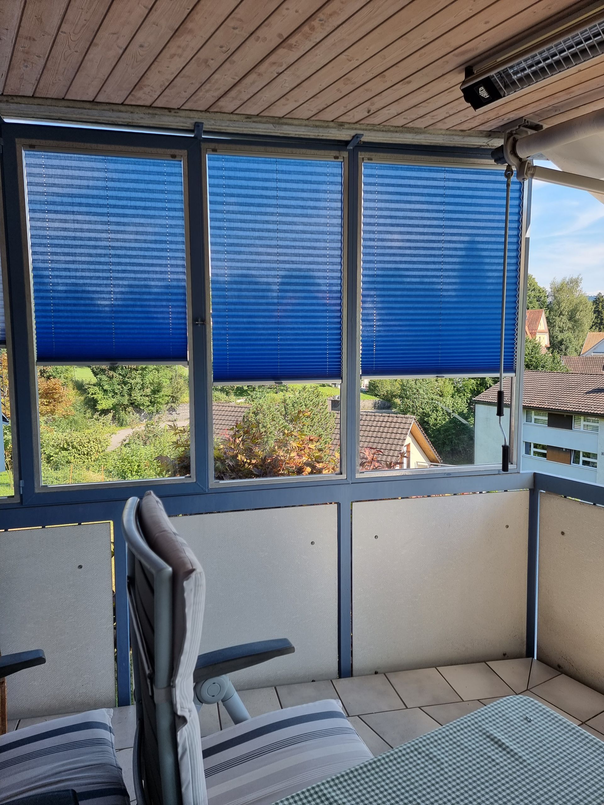 Blaue Plissees auf einem Balkon. Durch die Fenster sind ein Tisch, Stühle und ein Blick auf Bäume zu sehen.
