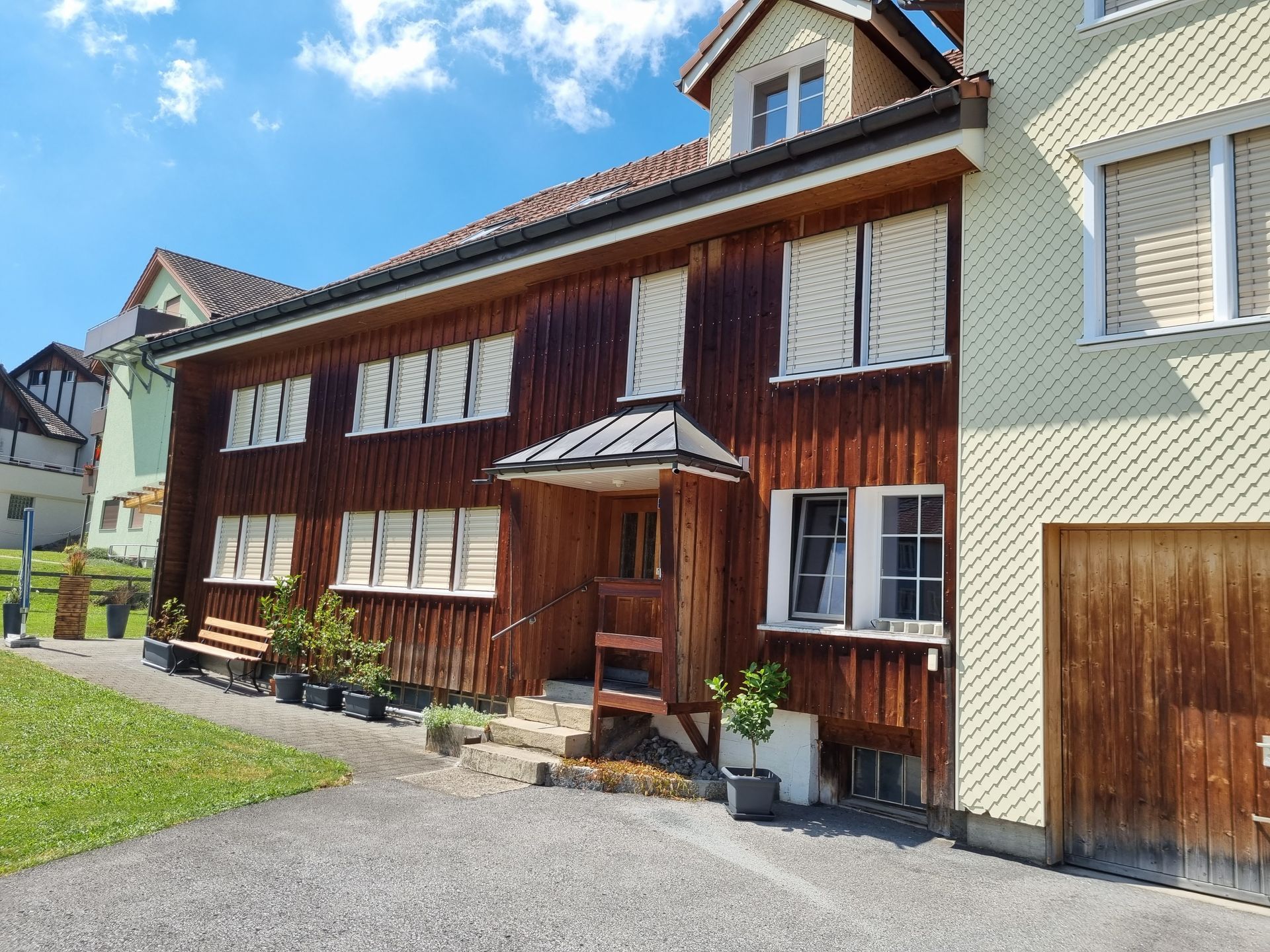 Holzhaus mit Fensterläden, Veranda und Stufen. Grüner Rasen, sonniger Tag.