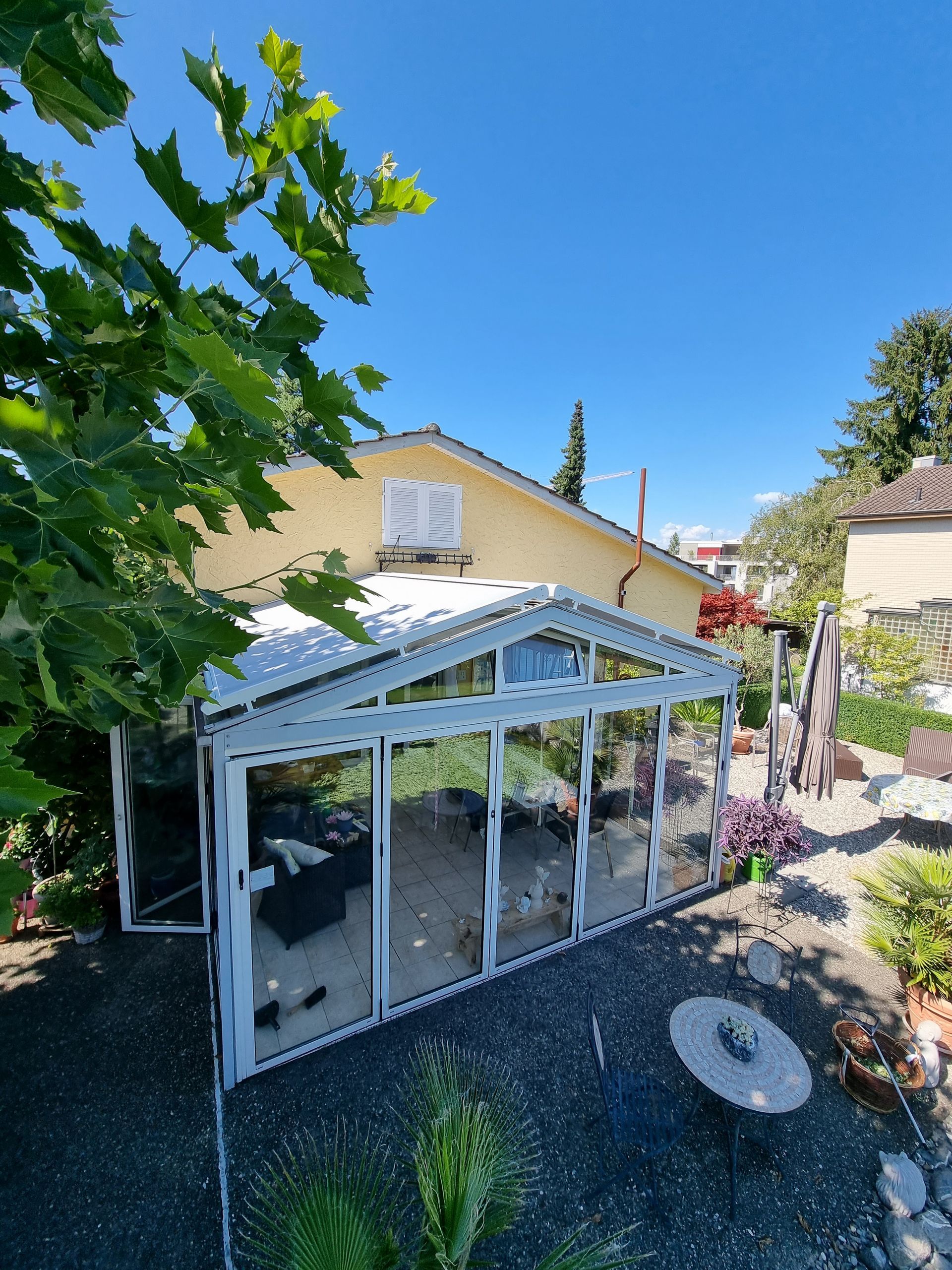 Ein Wintergarten mit Glaswänden an einem gelben Haus, sonniger Tag.