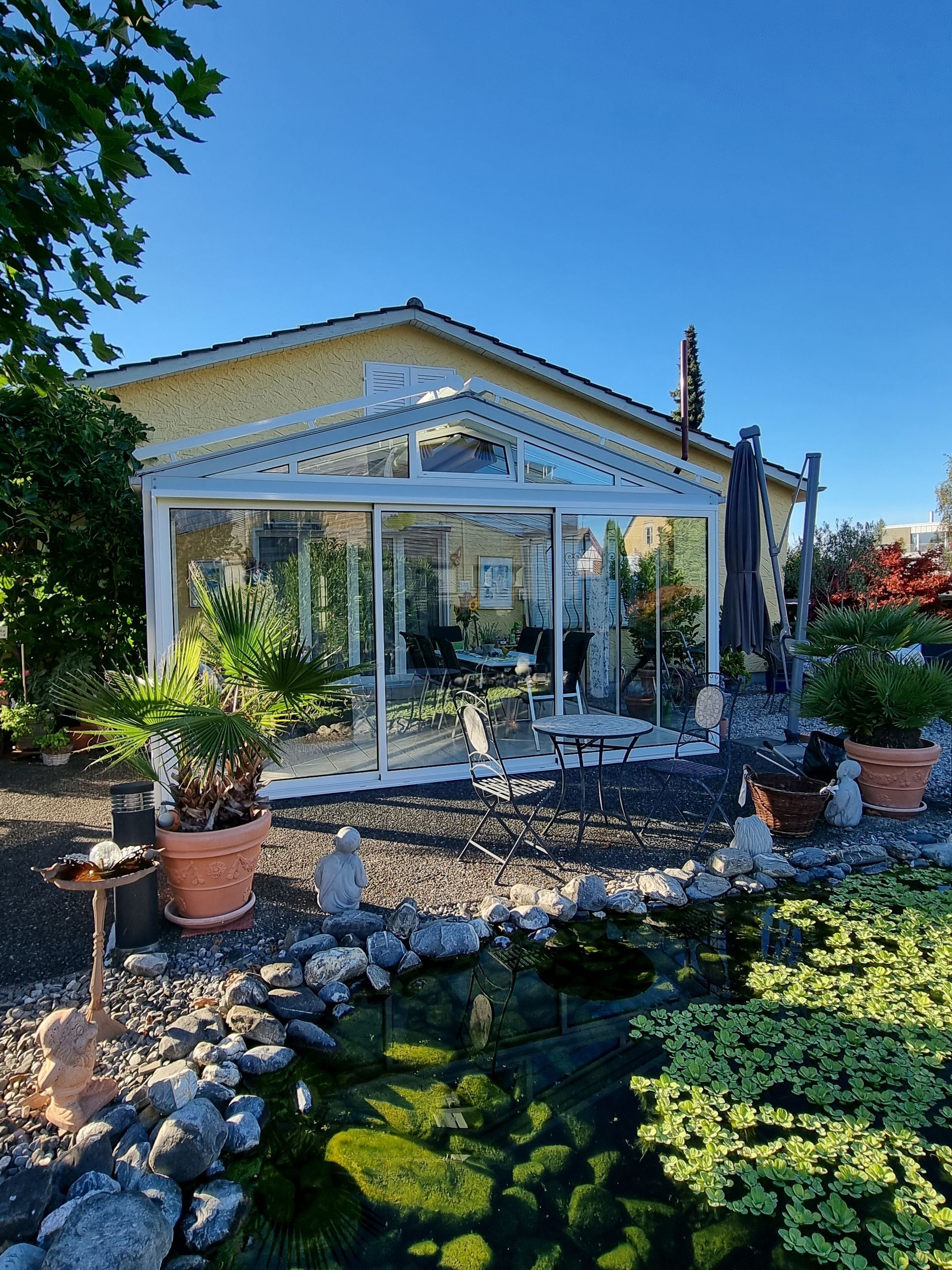 Gelbes Haus mit Wintergarten aus Glas, Teich, Topfpflanzen und klarem blauen Himmel.