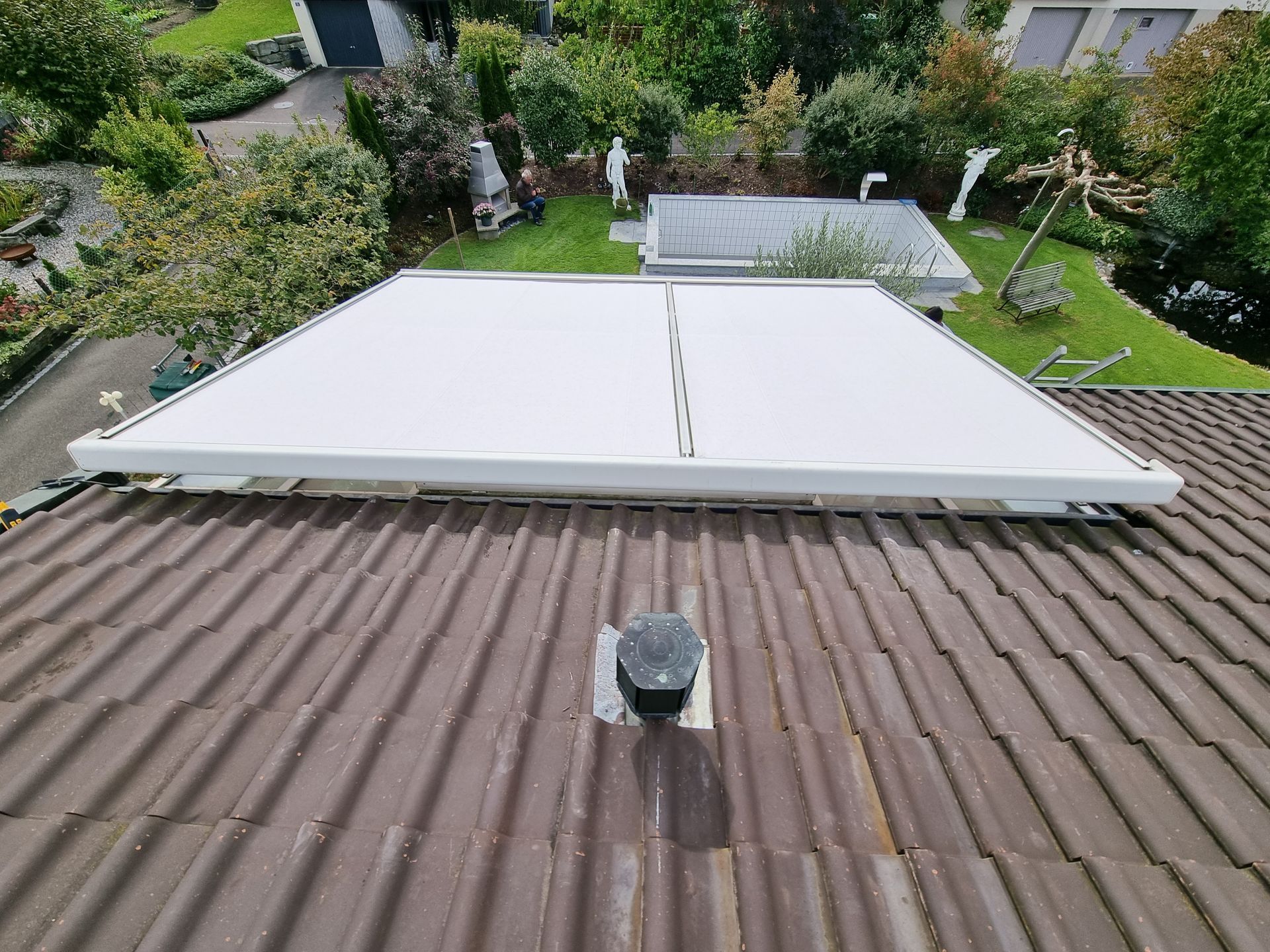 Weisse Markise auf einem braunen Ziegeldach. Blick von oben, mit grünen Bäumen und einer Wiese im Hintergrund.