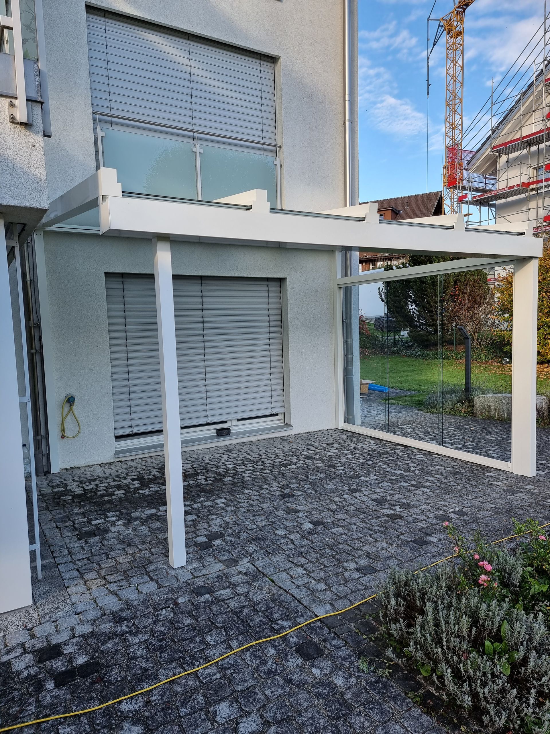 Terrassenüberdachung mit weissem Rahmen an einem weissen Gebäude mit geschlossenem Garagentor.