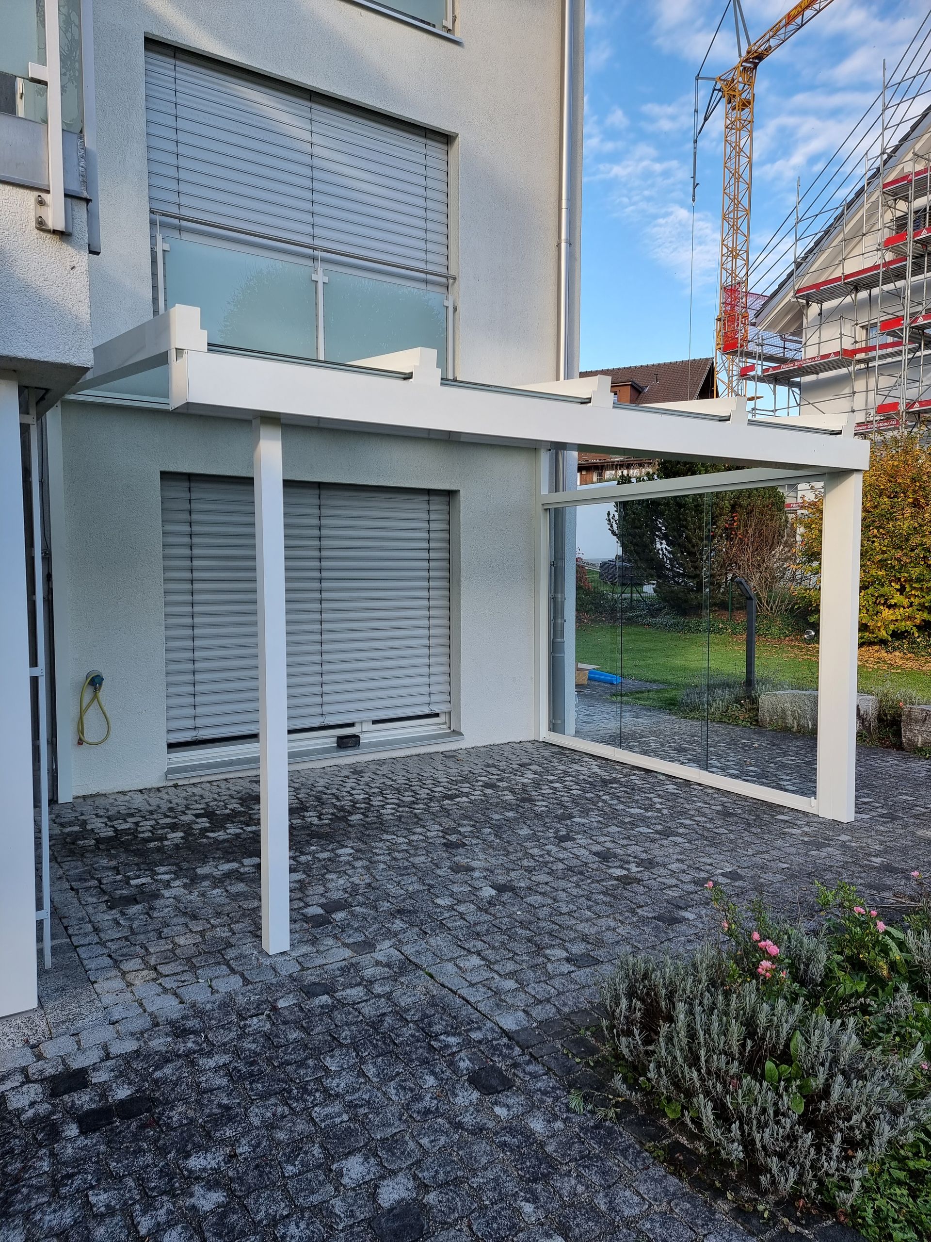 Weisse Pergola mit Glasdach und Glasseiten, befestigt an einem hellen Stuckgebäude, mit geschlossenen Jalousien.