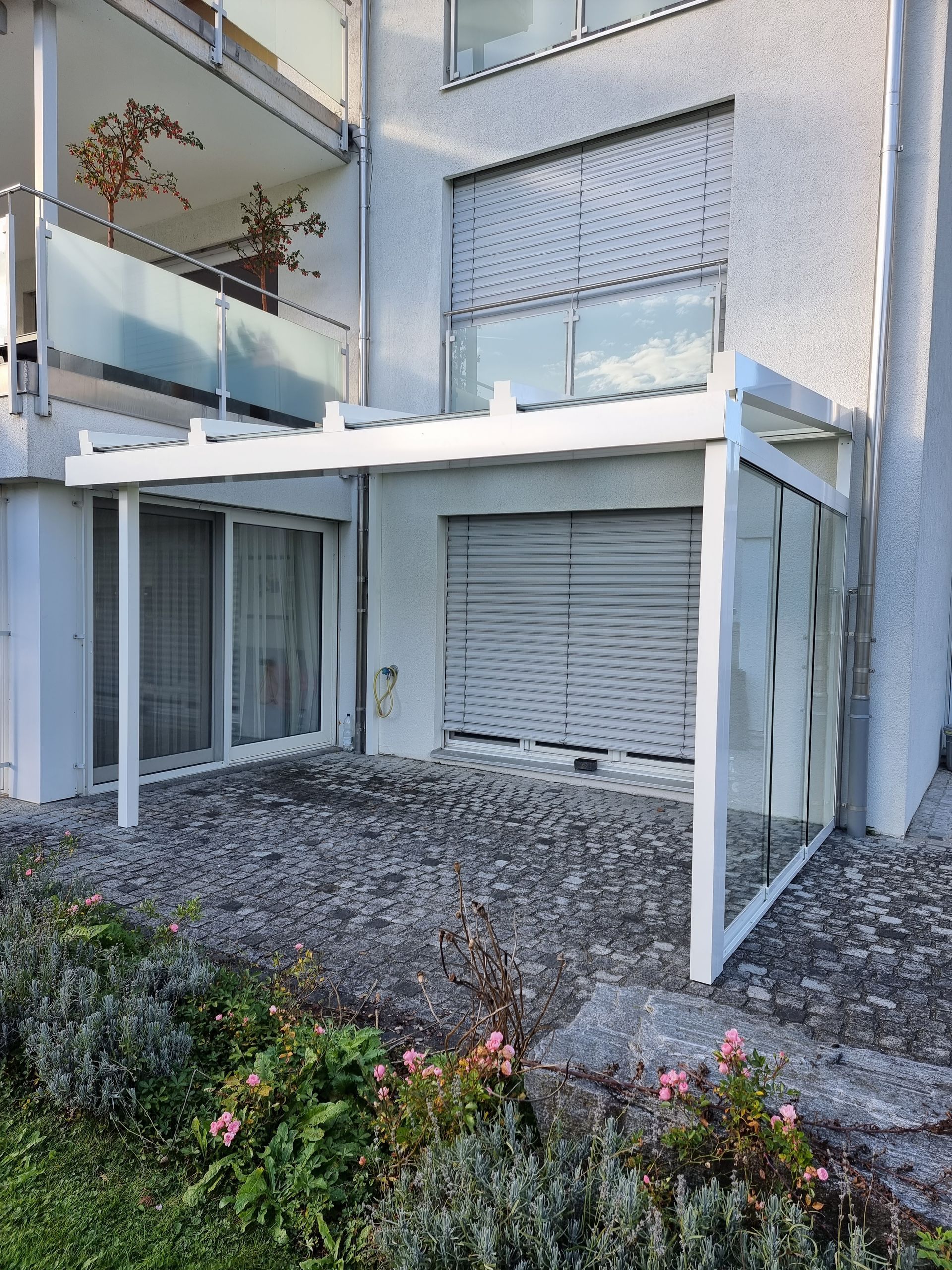 Weisse Terrassenüberdachung mit Glaswänden und -dach neben einem Gebäude mit Balkon.