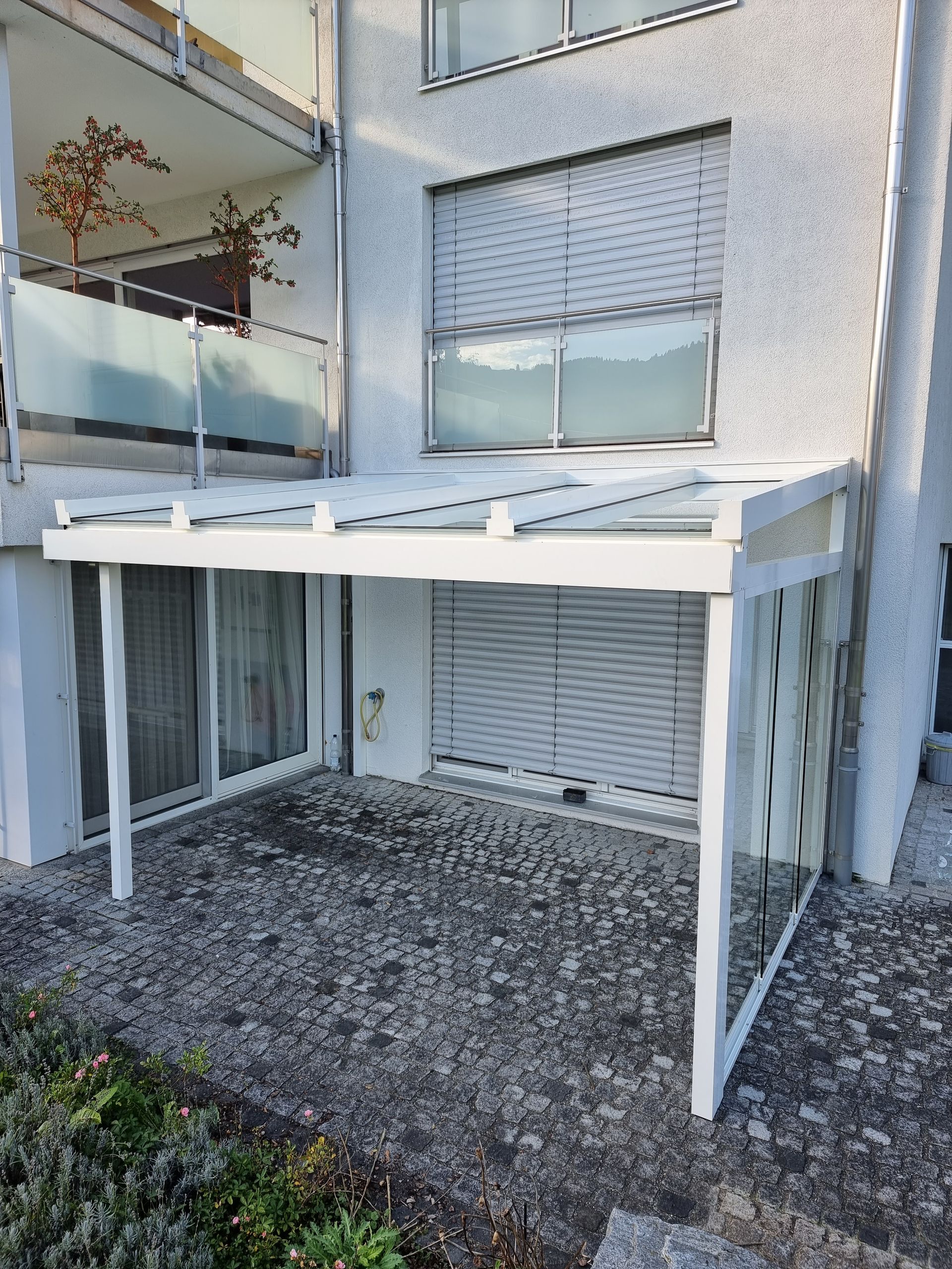An einem hellen Gebäude befestigte Terrassenüberdachung mit weissem Rahmen, geschlossenen Jalousien und Kiesboden.