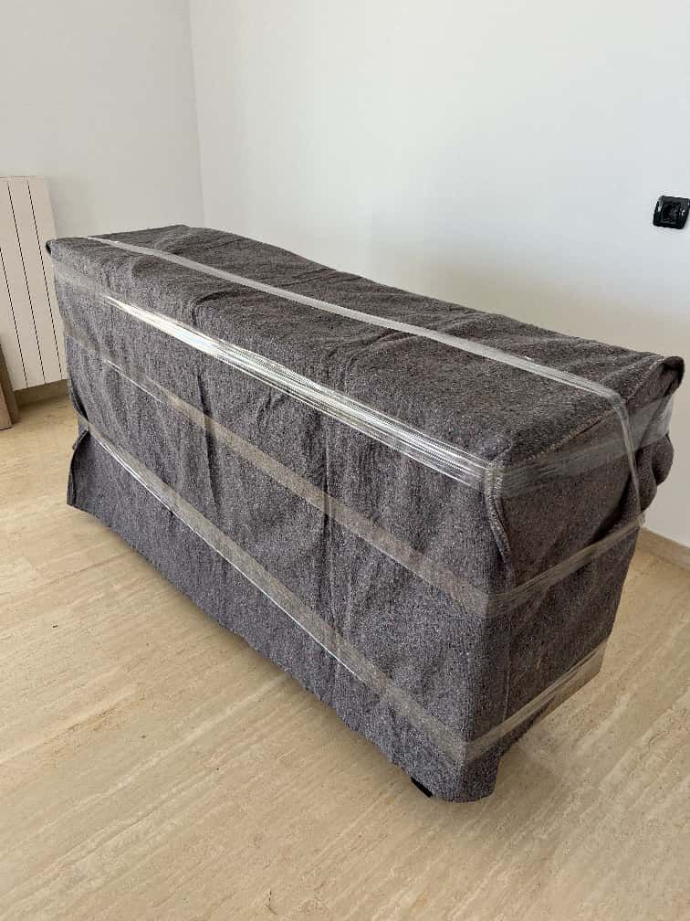 Un objeto grande y rectangular envuelto en una manta de mudanza gris, asegurado con cinta transparente, en el interior.