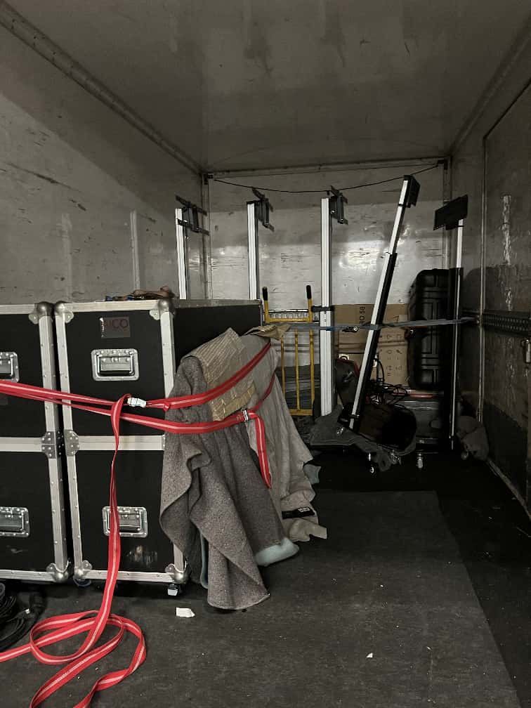 Dentro del área de carga de un camión, el equipo está sujeto con correas rojas. Se apilan cajas negras y otros equipos.