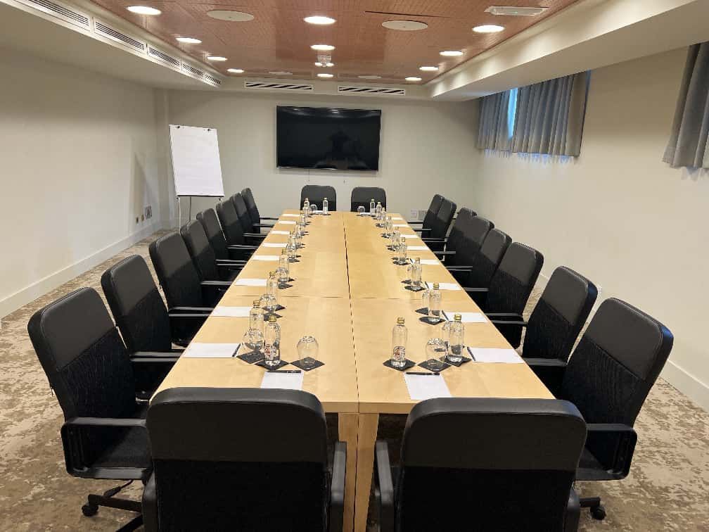 Sala de conferencias con una mesa larga, sillas negras y un televisor en la pared.
