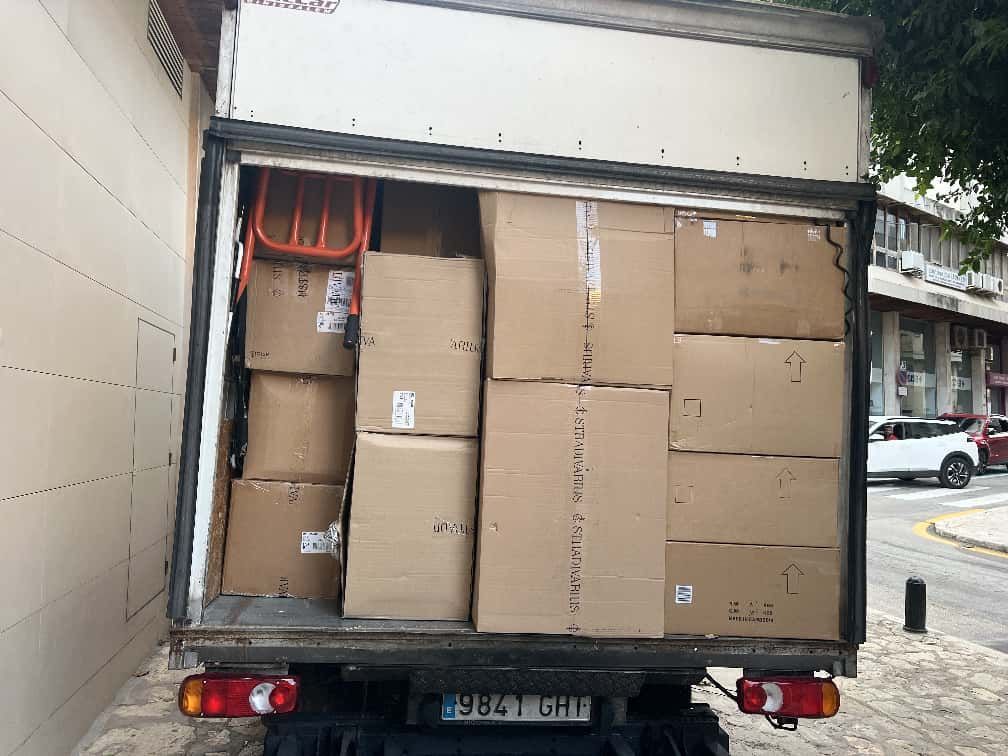 Vista trasera de un camión de reparto lleno de cajas de cartón, estacionado en una calle cerca de edificios.