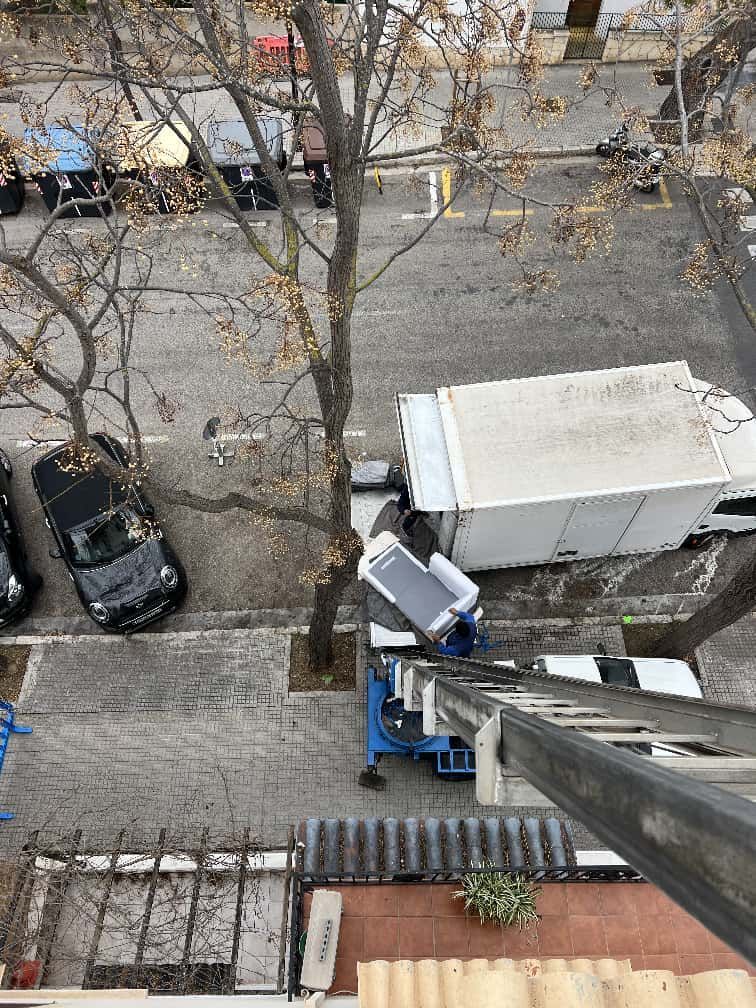 Camión de mudanzas con elevador descargando muebles en una calle de la ciudad. Pavimento gris, árboles desnudos y coches.