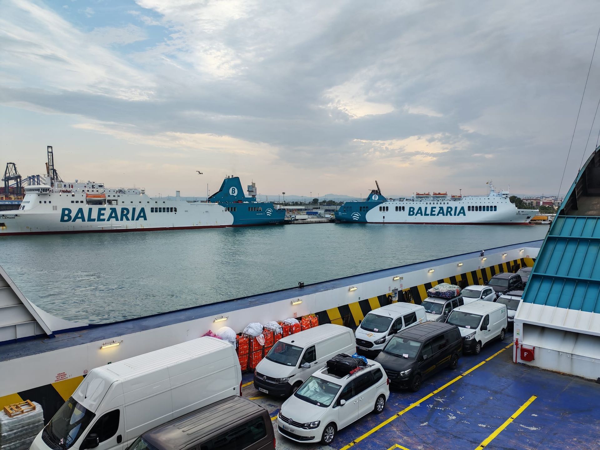 Automóviles y camiones en la cubierta de un ferry; otros dos ferrys de Baleària atracados en el puerto bajo un cielo nublado.