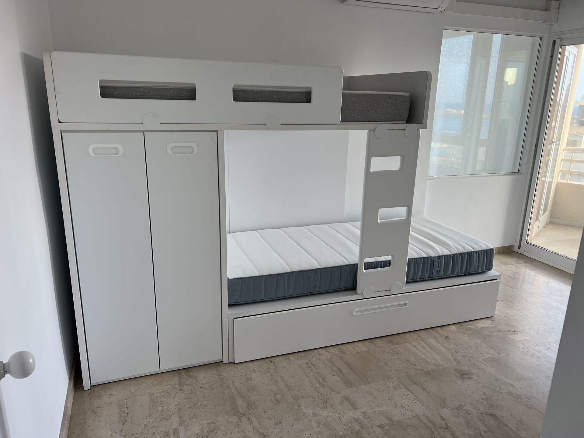 Litera blanca con armario, cajones y colchón, en habitación con ventana.