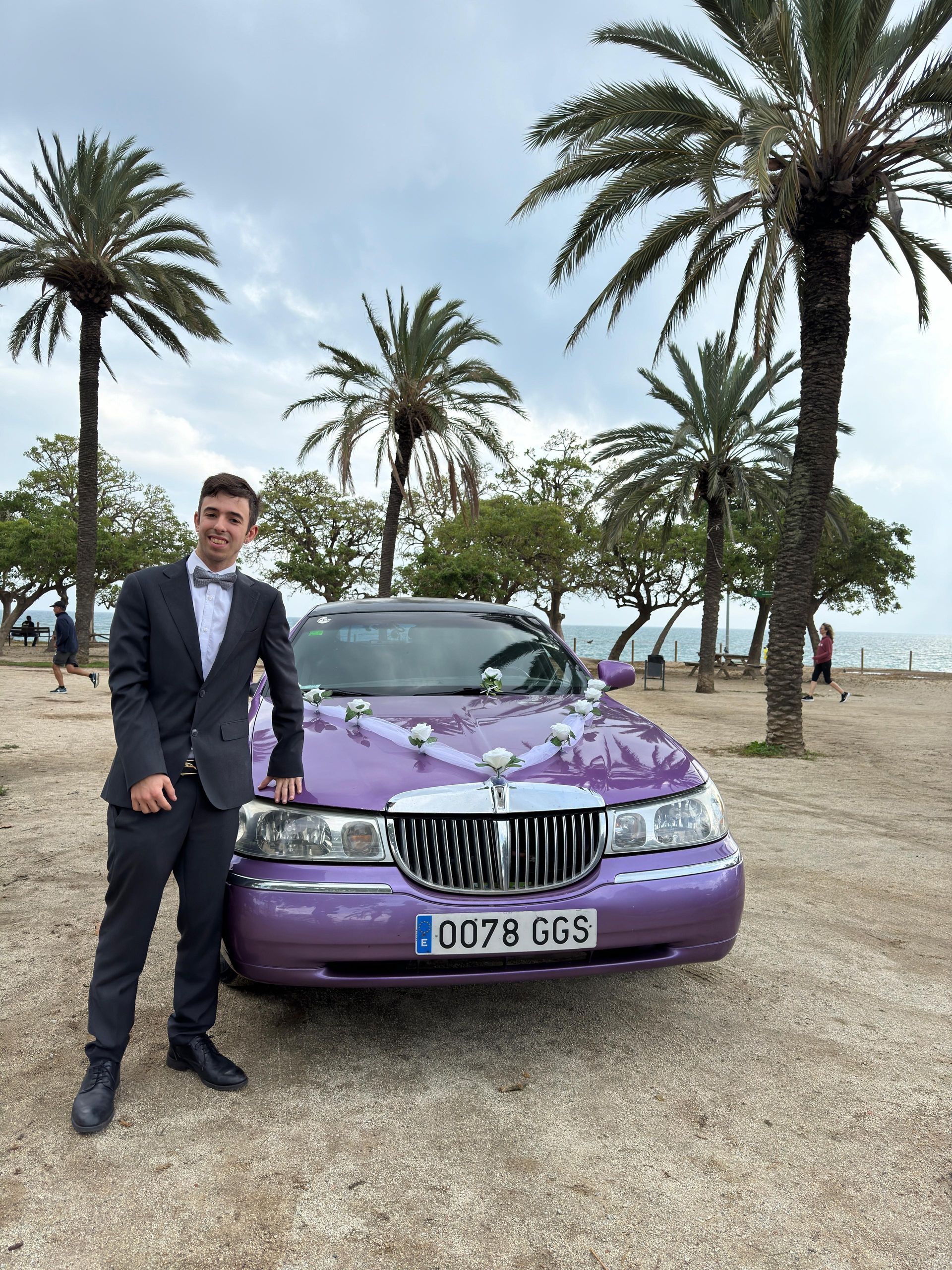 Un hombre con traje y pajarita está de pie junto a una limusina violeta.