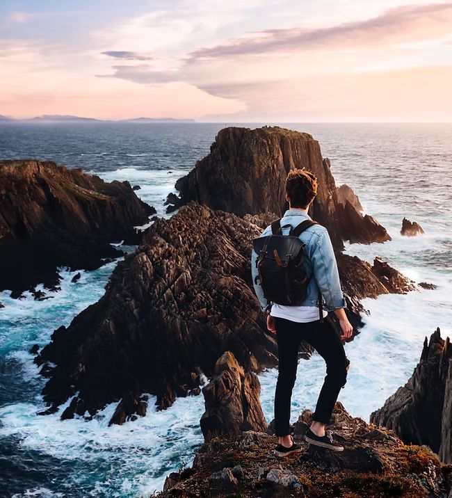 A man with a backpack is standing on top of a rocky cliff overlooking the ocean.