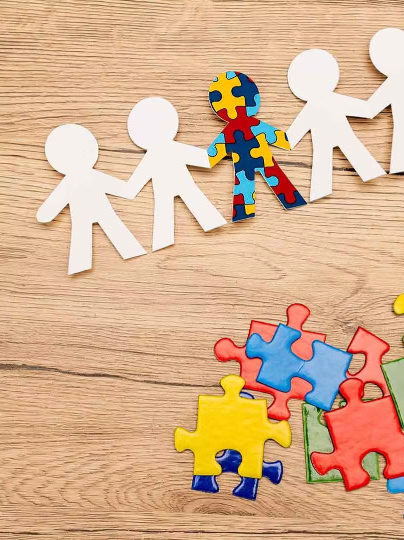 A group of paper people holding hands next to puzzle pieces on a wooden table.