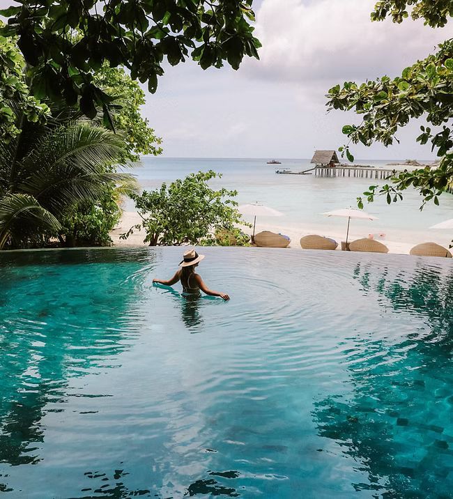 A woman in a hat is swimming in an infinity pool overlooking the ocean
