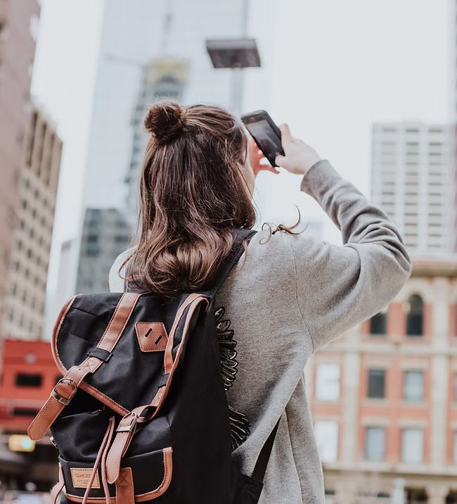 A woman with a backpack is taking a picture of a city with her phone.