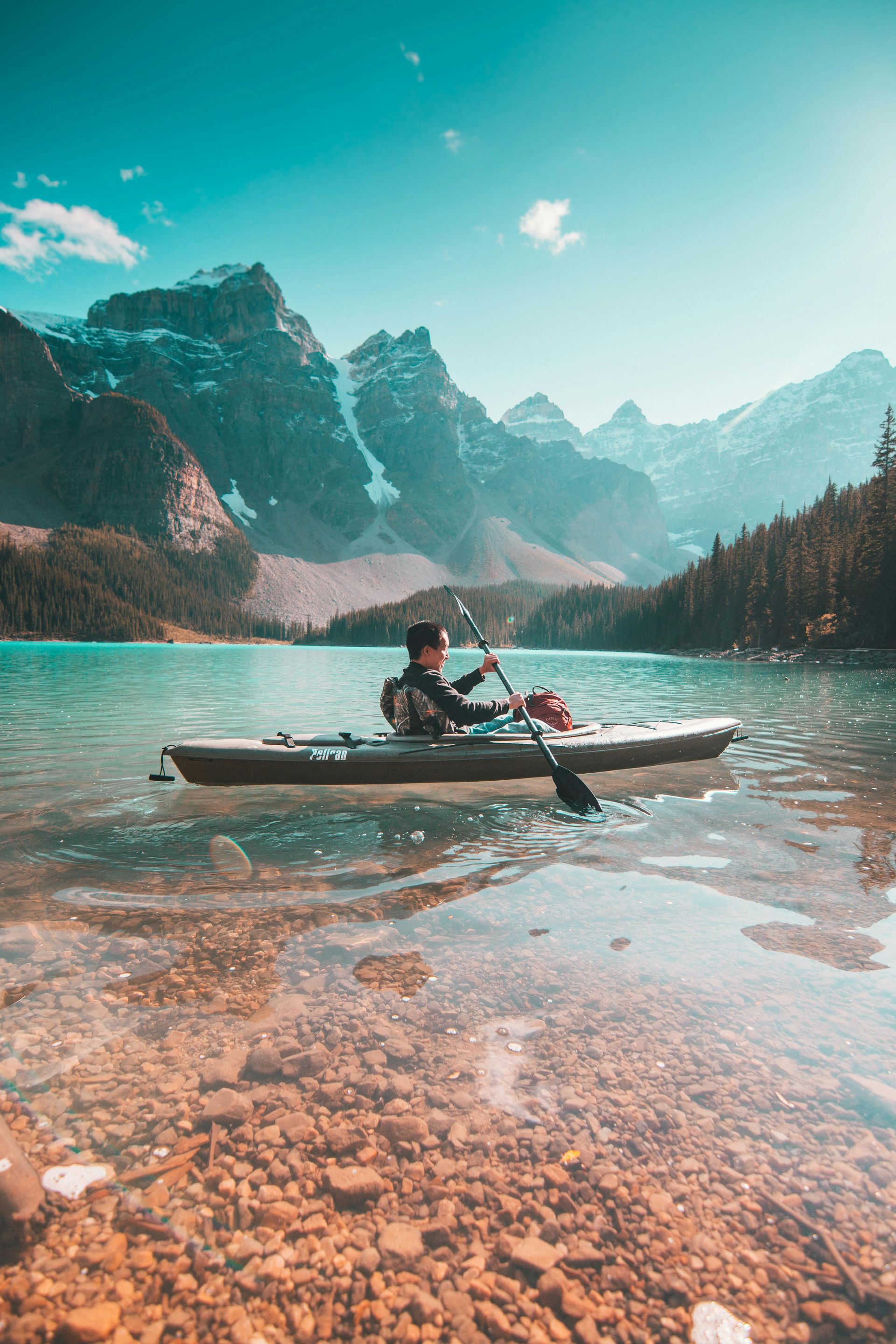 A man is paddling a kayak on a lake with mountains in the background.